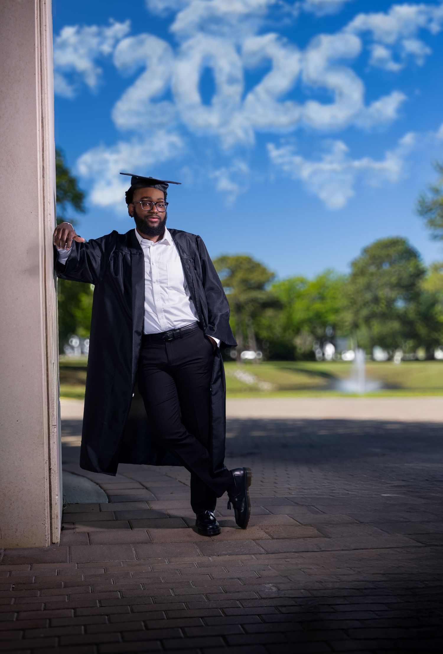 Graduate posing against a pillar with '2025' written in clouds above in a celebratory graduation photo. Portrava Creative Studios