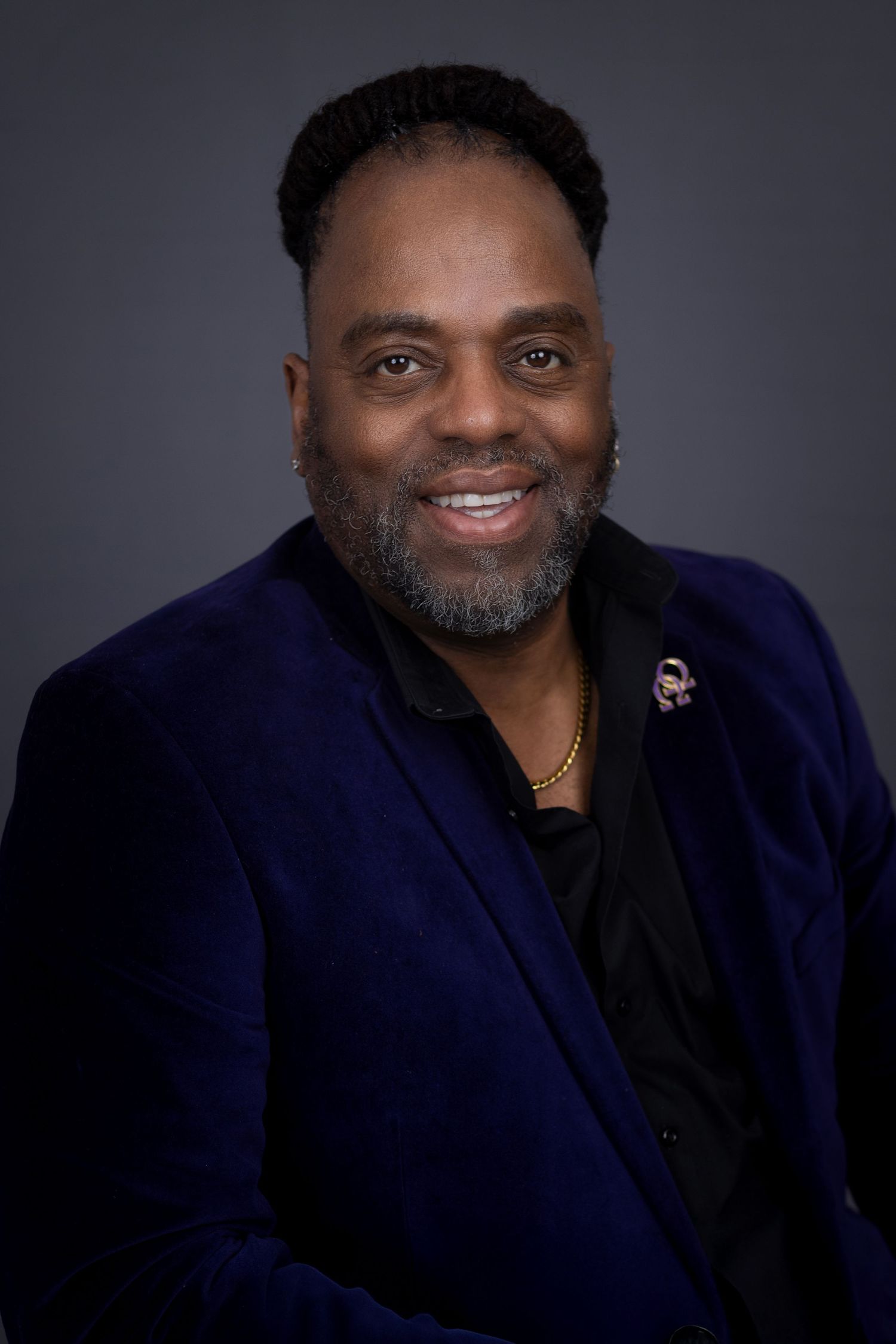 Professional headshot of a person in a navy velvet blazer and black shirt smiling warmly against a dark background. Portrava Creative Studio