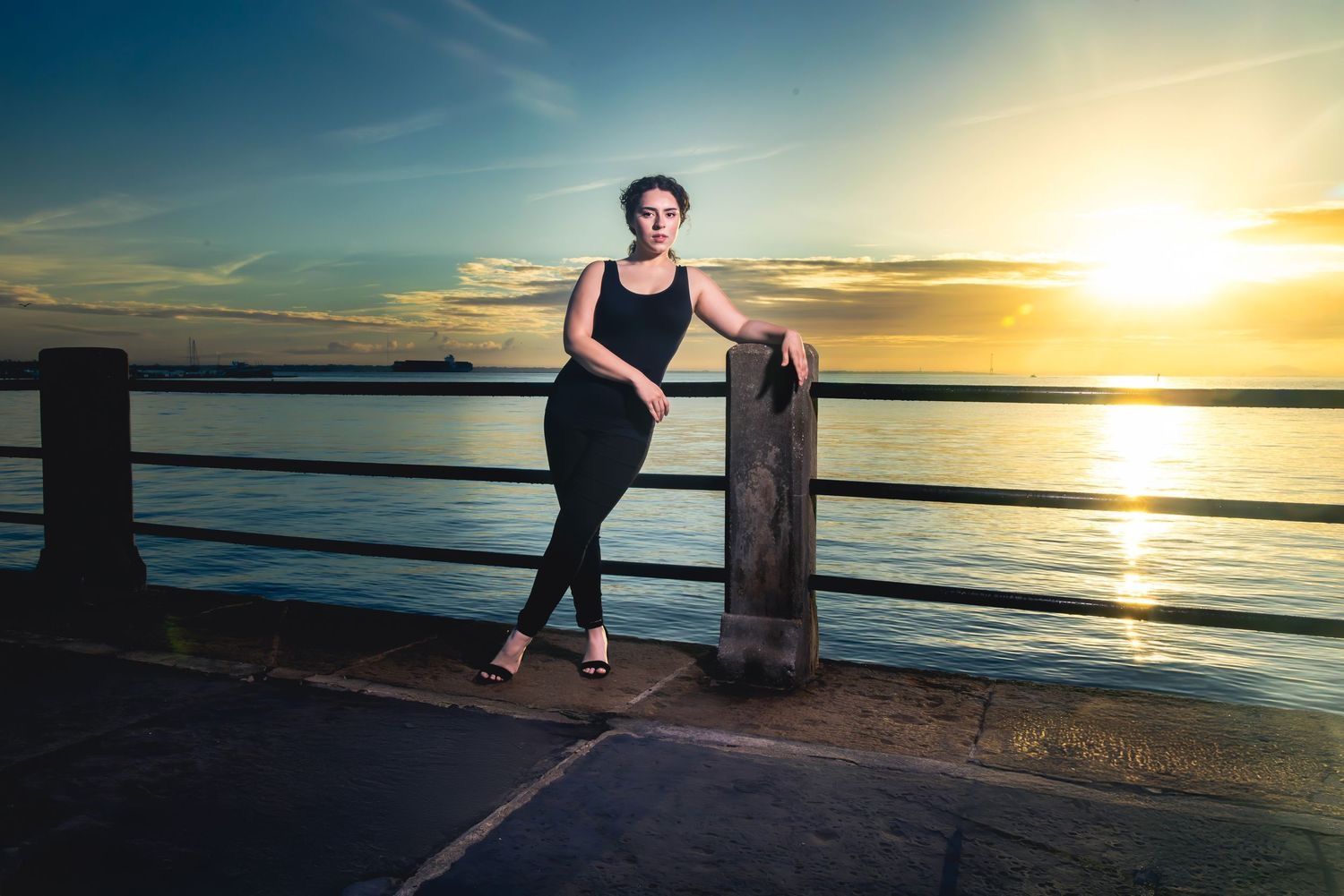 A silhouetted figure poses against a vibrant sunset over water on a pier. Portrava Creative Studios