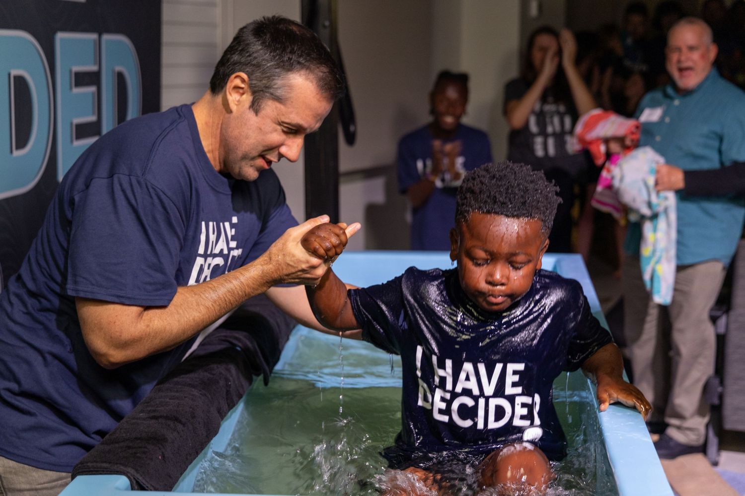 Young child in baptismal pool wearing 'I Have Decided' t-shirt during church ceremony. Portrava Creative Studios