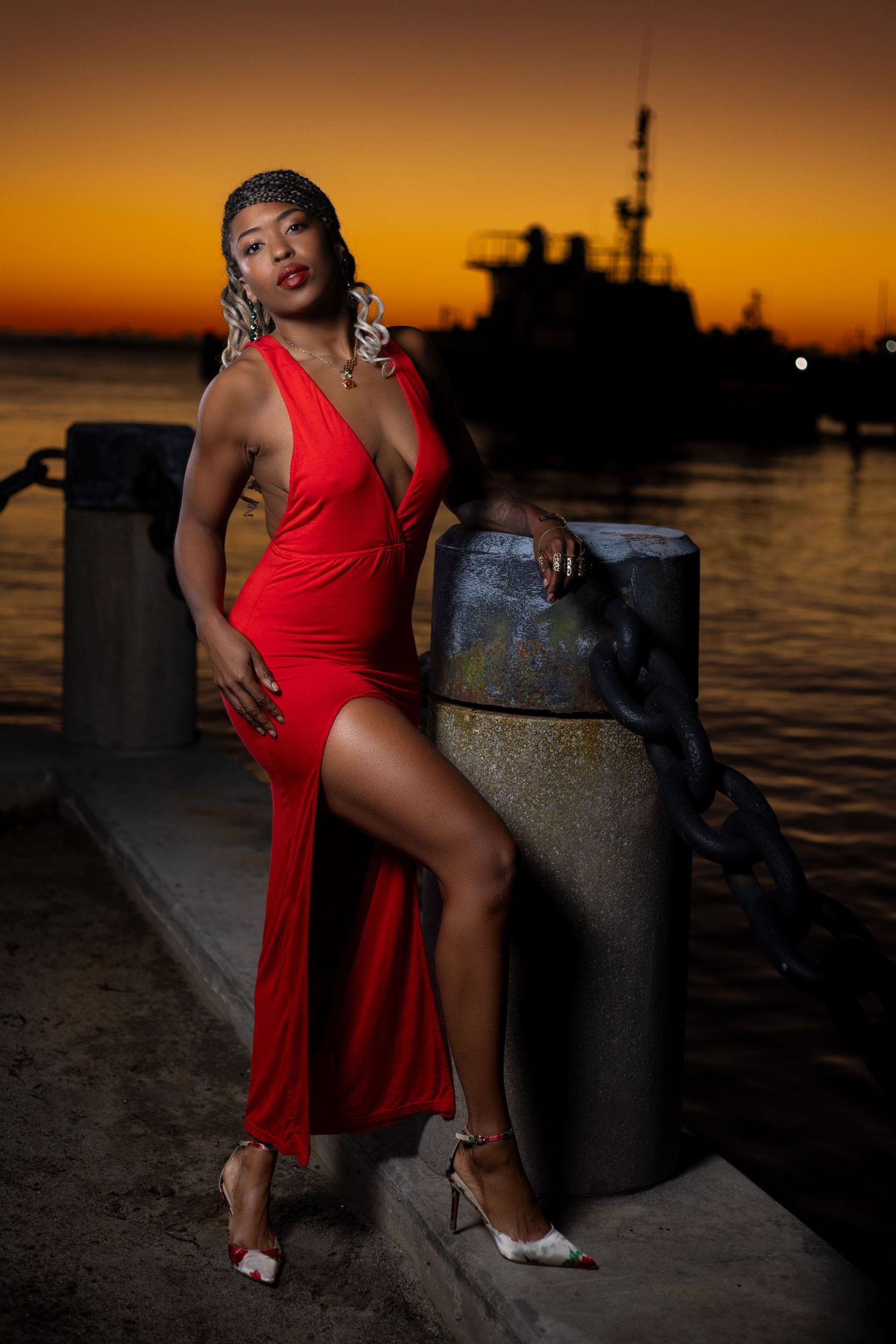Dramatic sunset portrait of figure in red dress by harbor with ship silhouette. Portrava Creative Studios