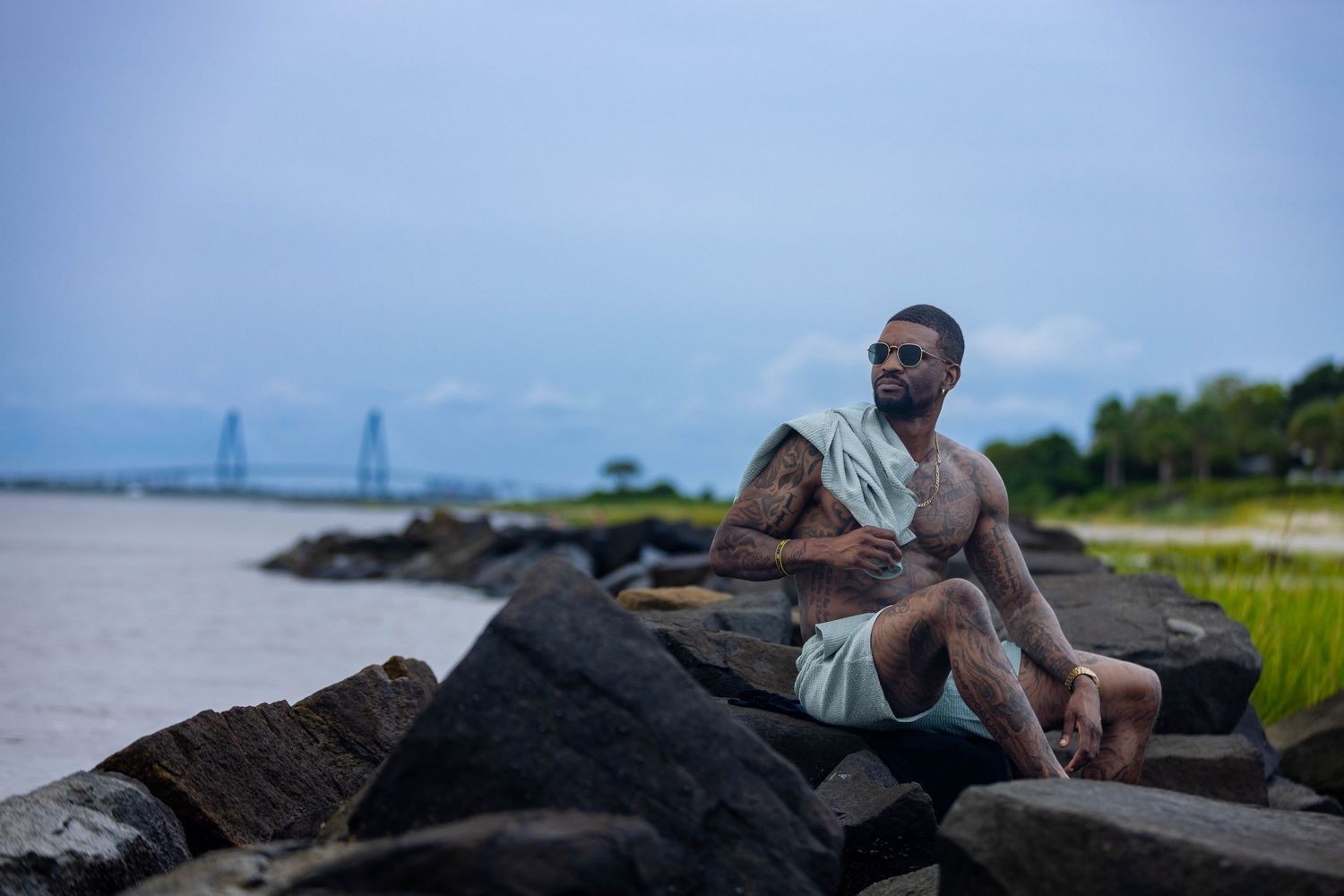 Person relaxing on rocky shoreline with water and bridge in background. Portrava Creative Studios