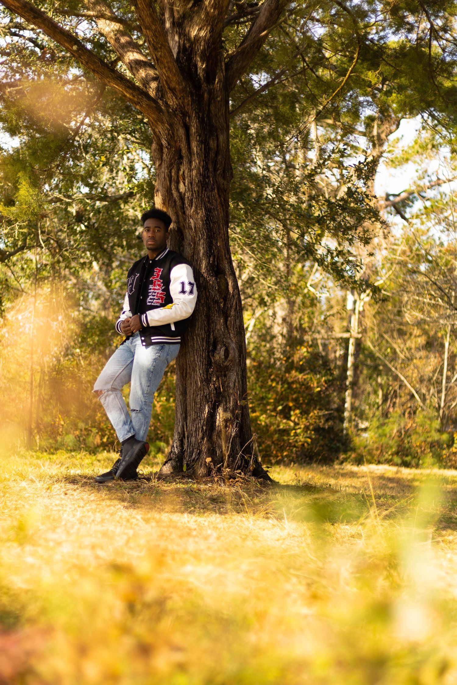 Person in varsity jacket leans against large tree in golden sunlit field. Portrava Creative Studios