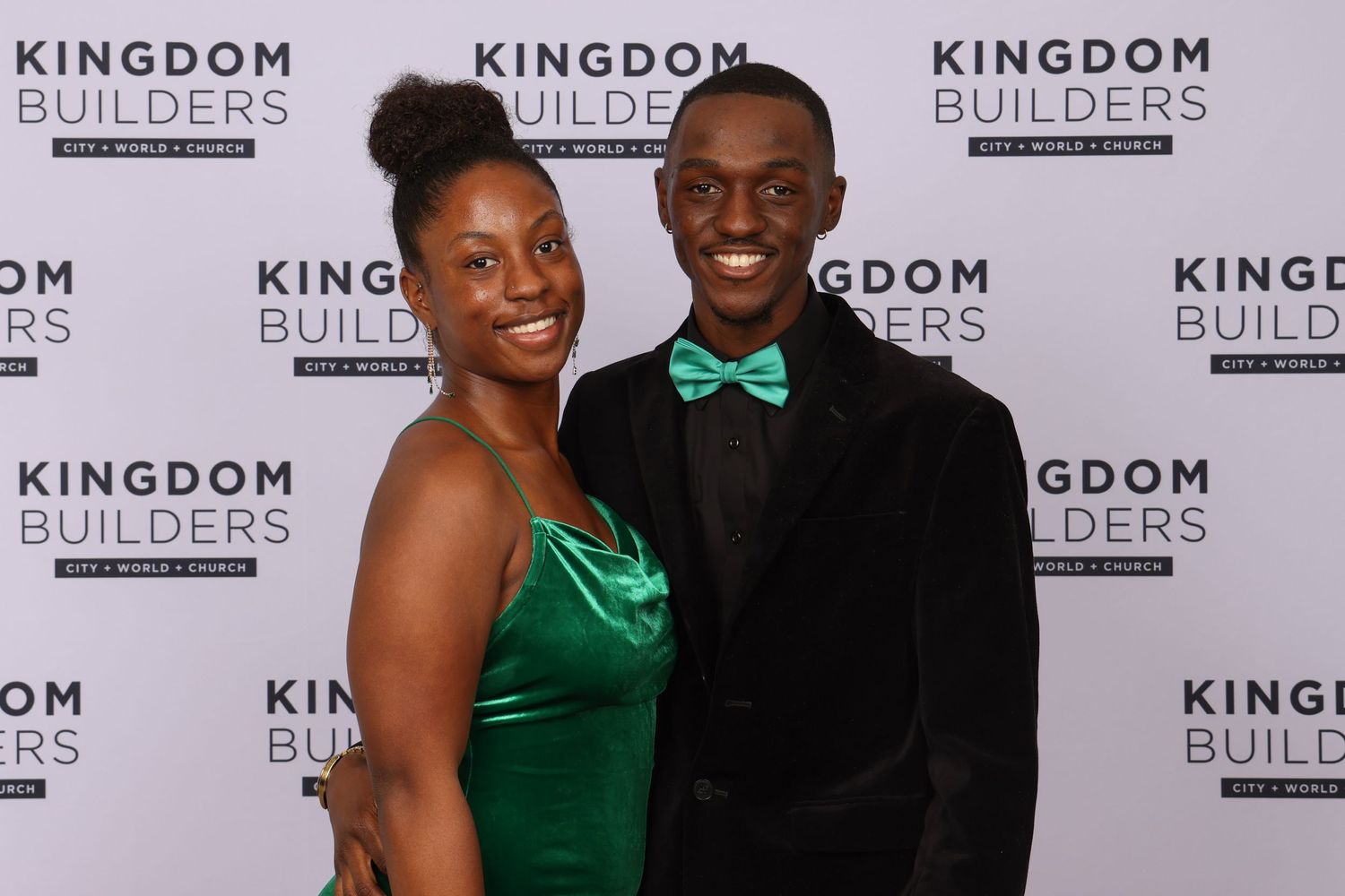 Anthony Cummings Jr and his Fiance in formal attire poses at Kingdom Builders event backdrop, with green dress and bowtie accents. Portrava
