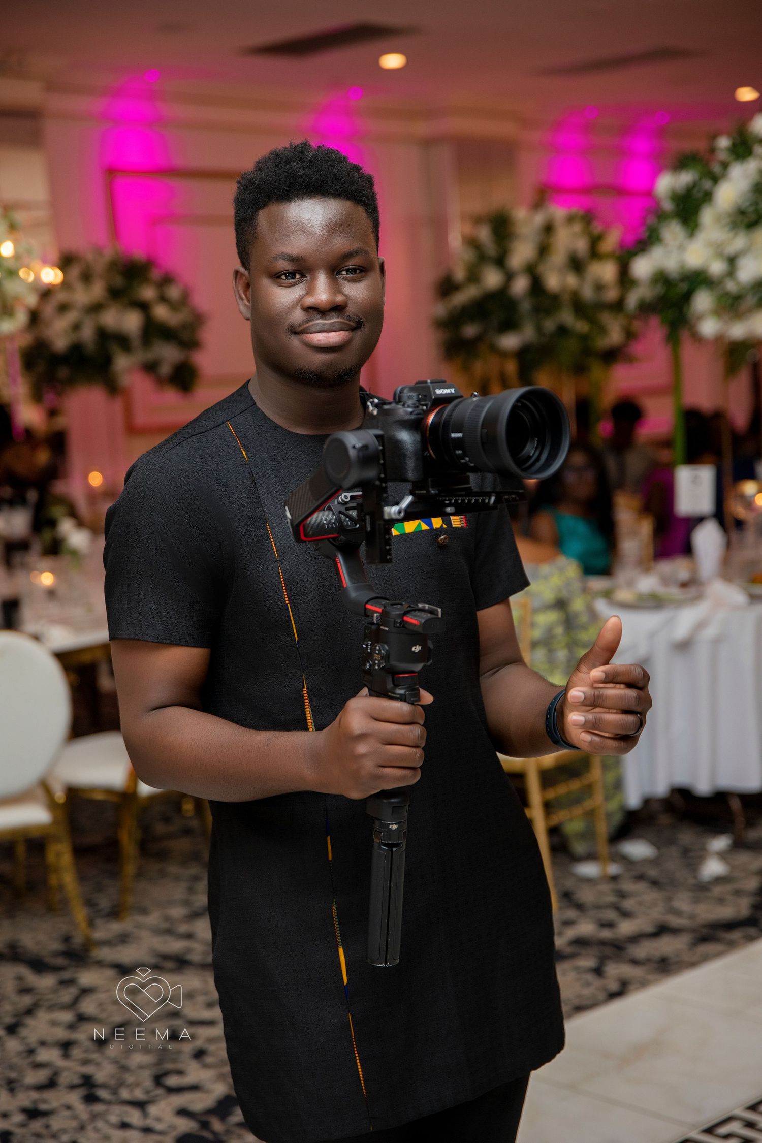 Videographer holding professional camera equipment at an elegant event with pink lighting and floral decor.