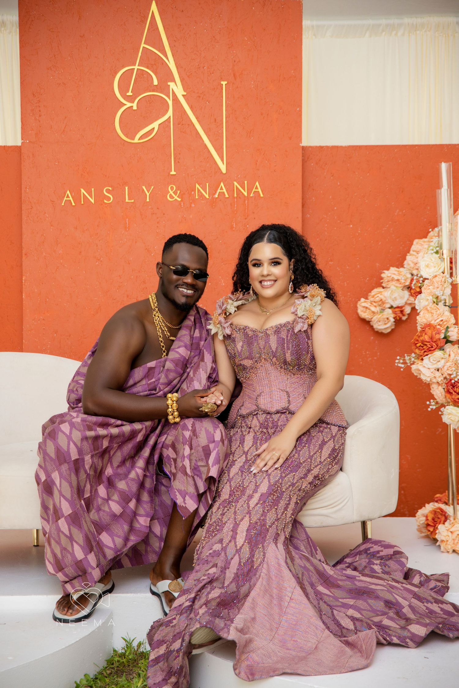 Two people in matching pink gowns pose against a coral wall with gold logo signage.