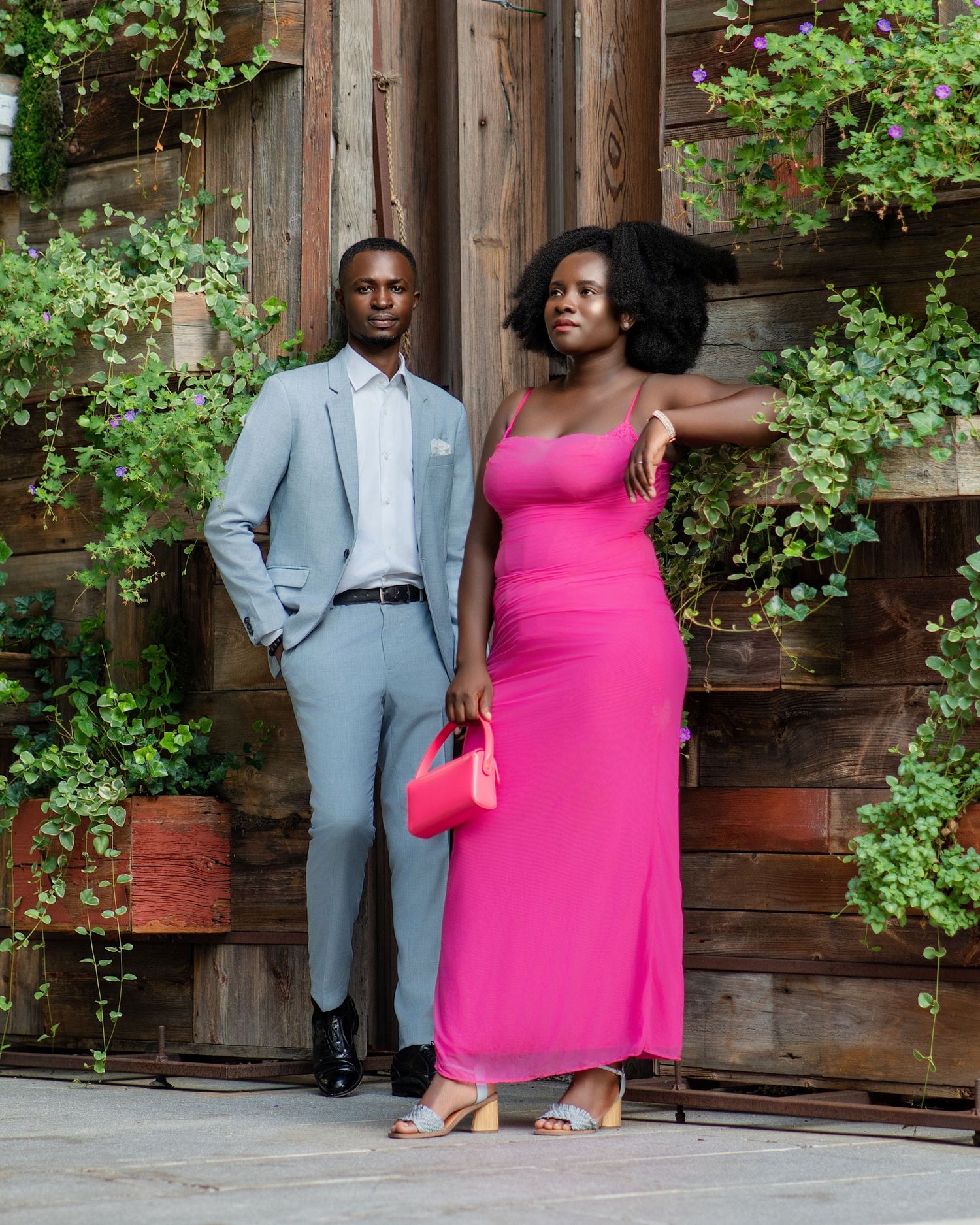 Fashion-forward couple in bright pink dress and light blue suit pose by wooden wall with greenery.