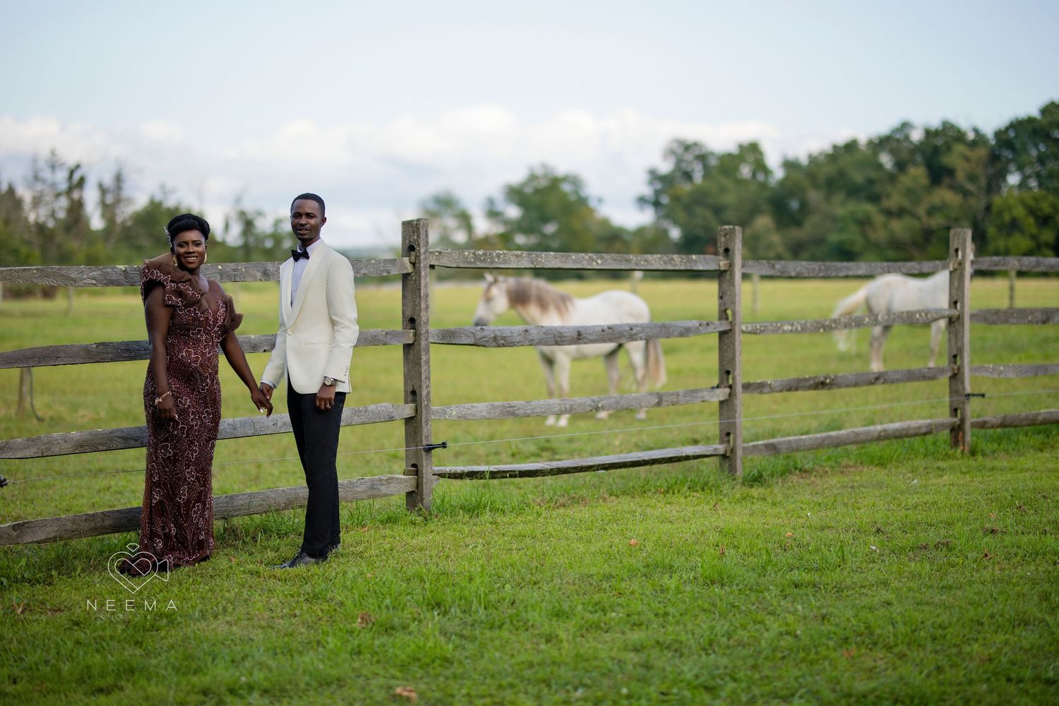 Couple standing near rustic wooden fence with horses grazing in background.