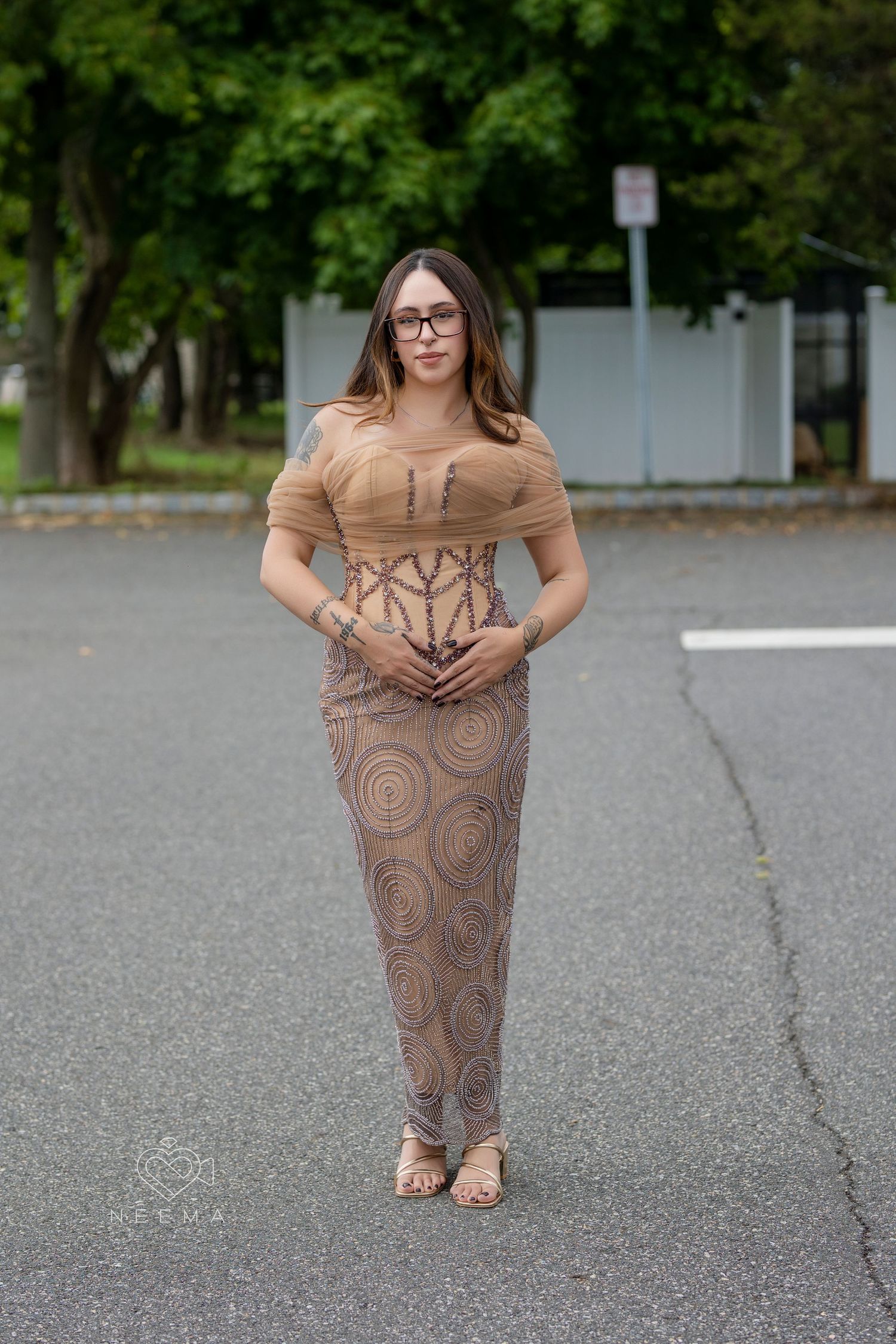 Woman in tan and beige patterned dress poses in parking lot with trees in background.