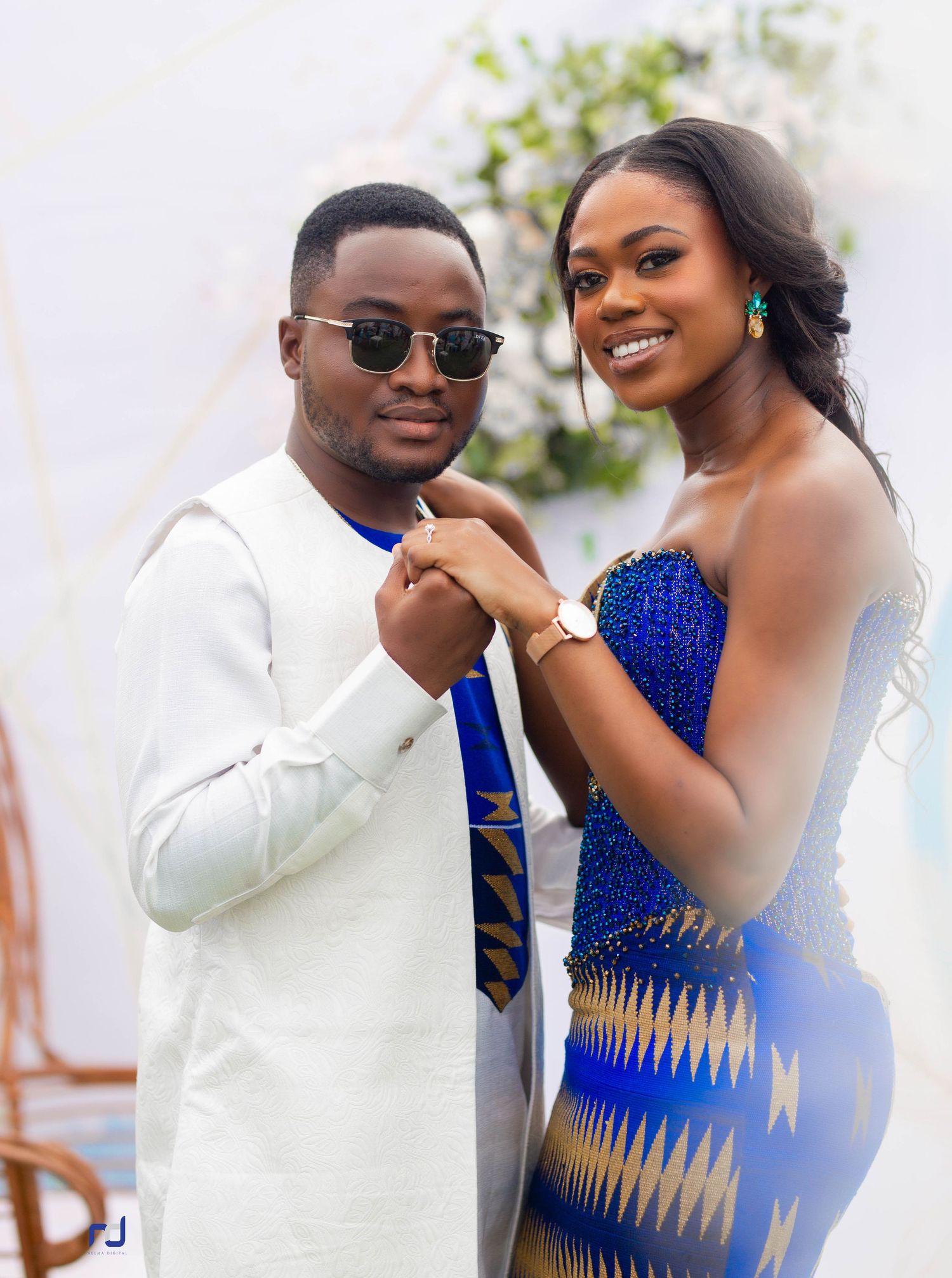 A couple poses in formal wear, white suit and royal blue beaded dress at an outdoor event.