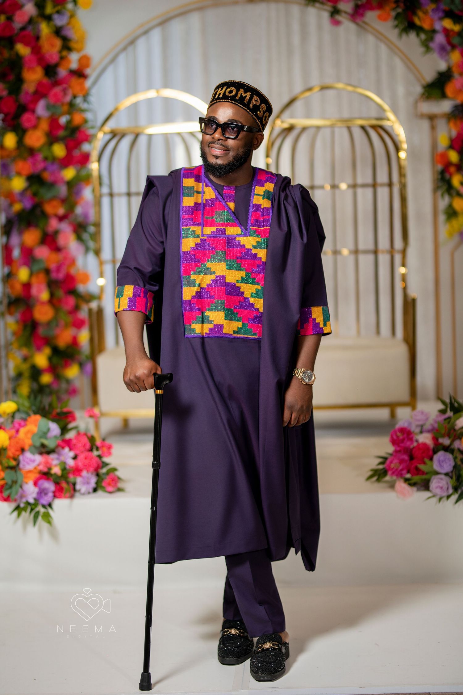 A person in a purple traditional African outfit with colorful geometric patterns poses with a walking stick.