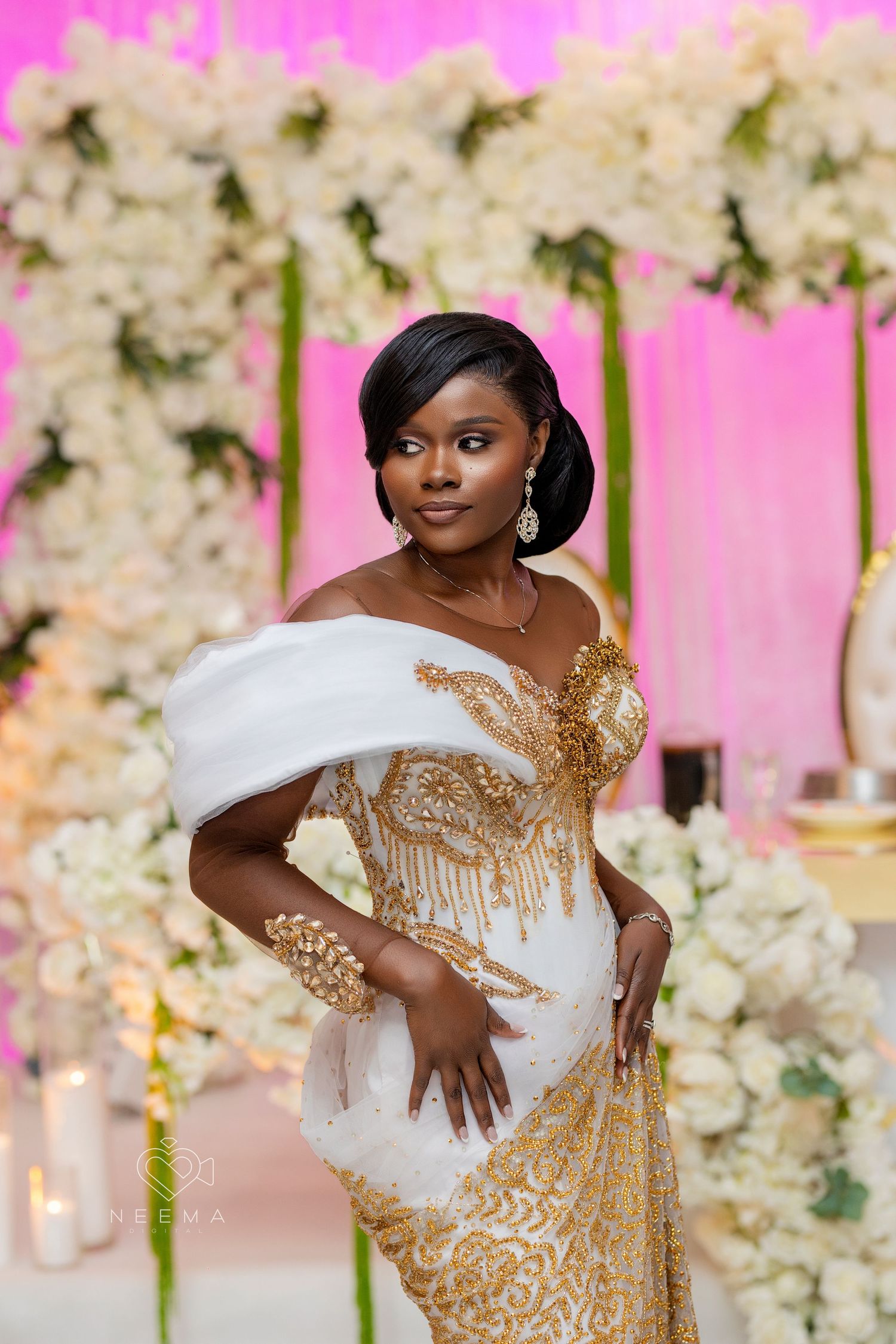 Bride in stunning white and gold dress poses against floral backdrop with pink lighting.