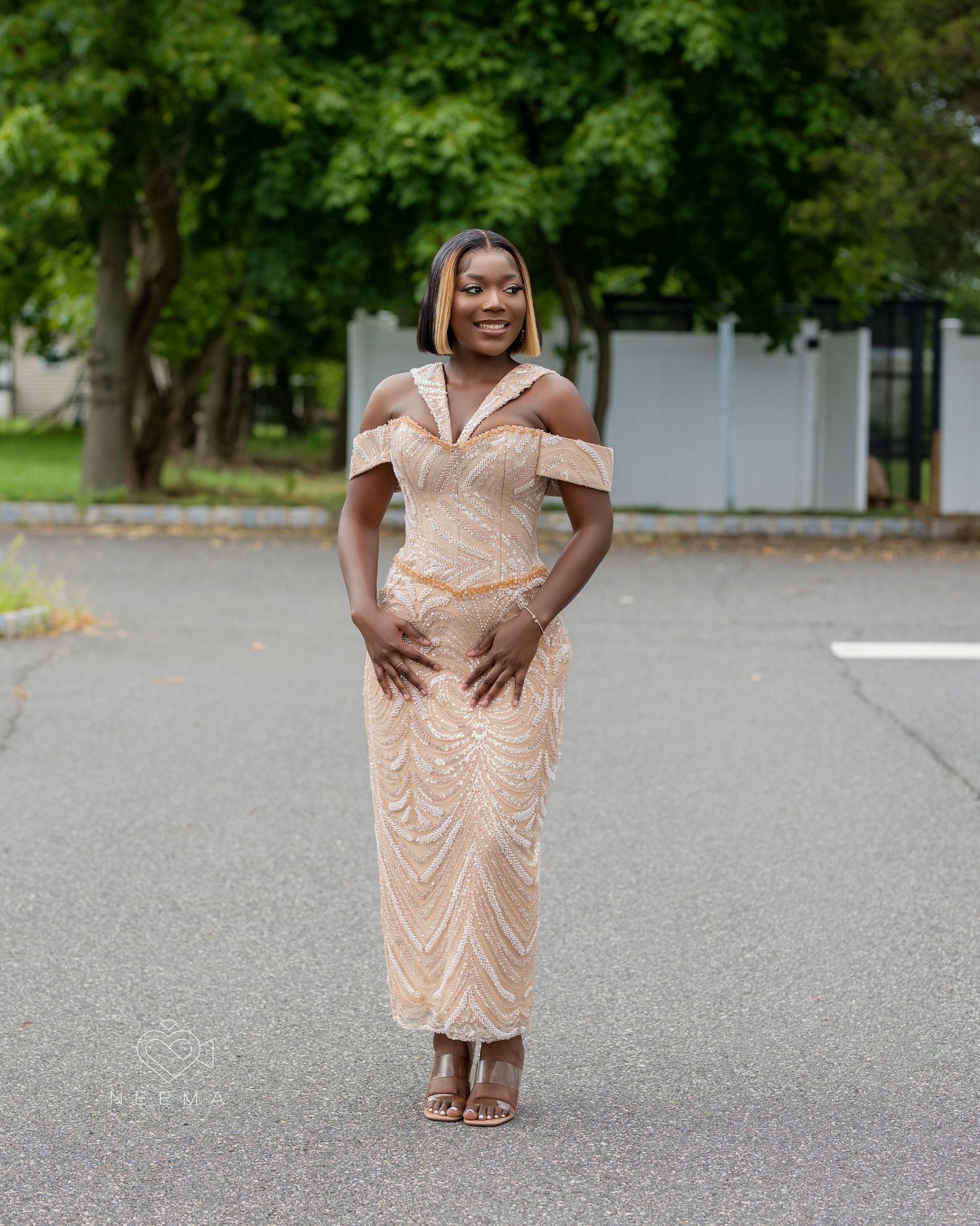 Woman wearing off-shoulder peach gown poses elegantly in parking area.