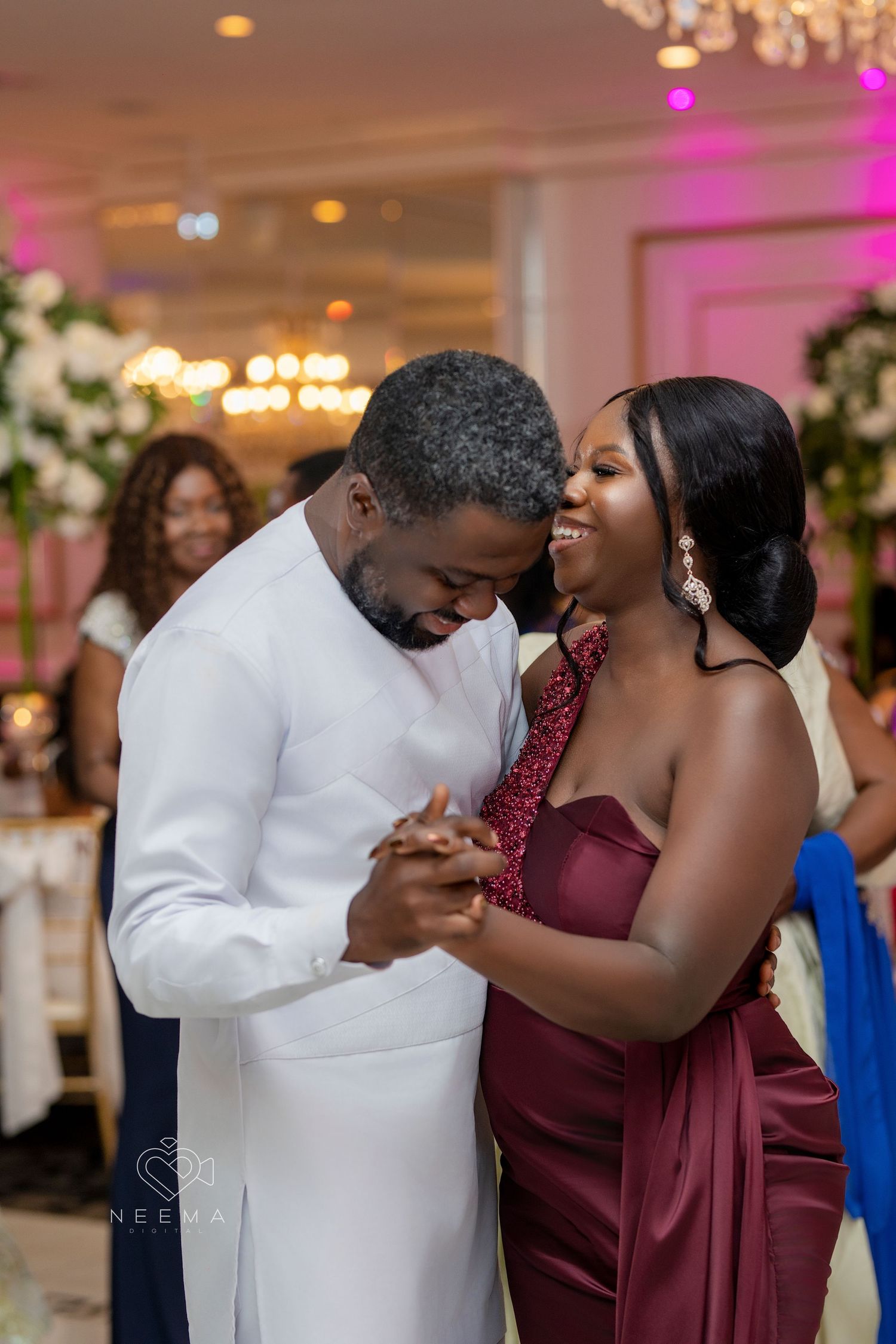 A couple dancing together at an elegant reception with pink lighting and white floral decorations.