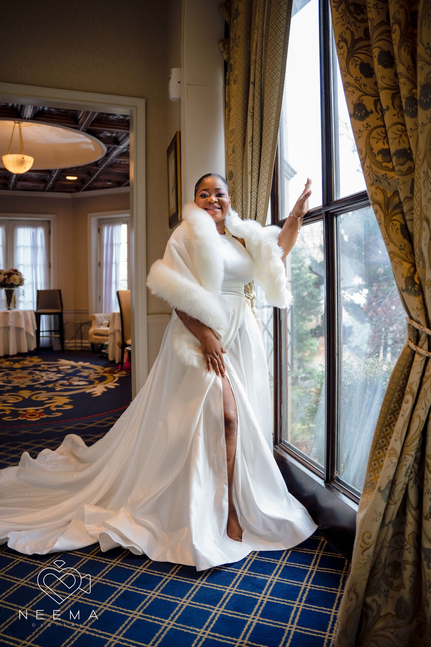 Bride in flowing white gown and fur stole poses dramatically by ornate window.