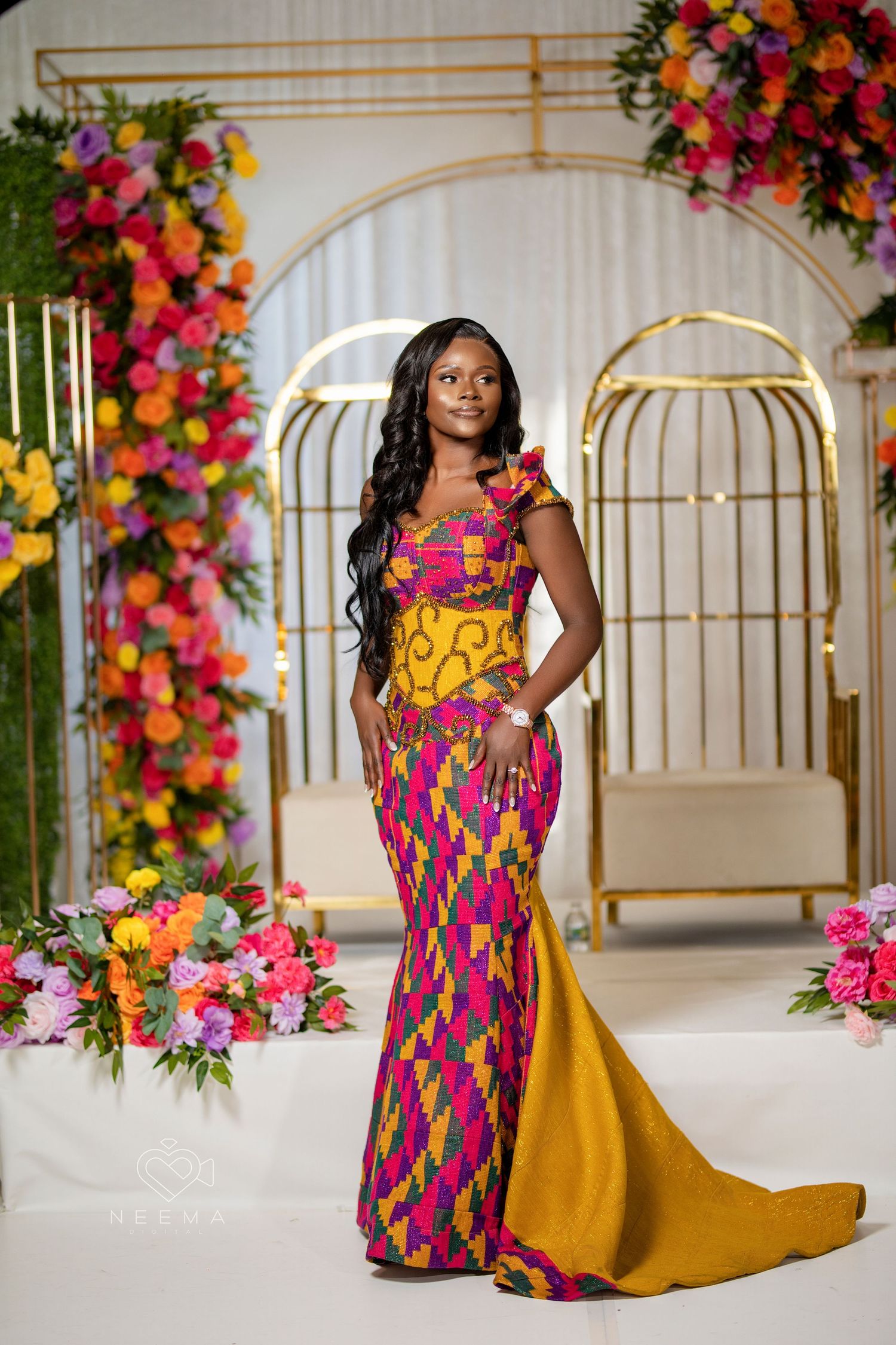 A woman in a vibrant African print gown poses against a floral backdrop with golden bird cage decorations.