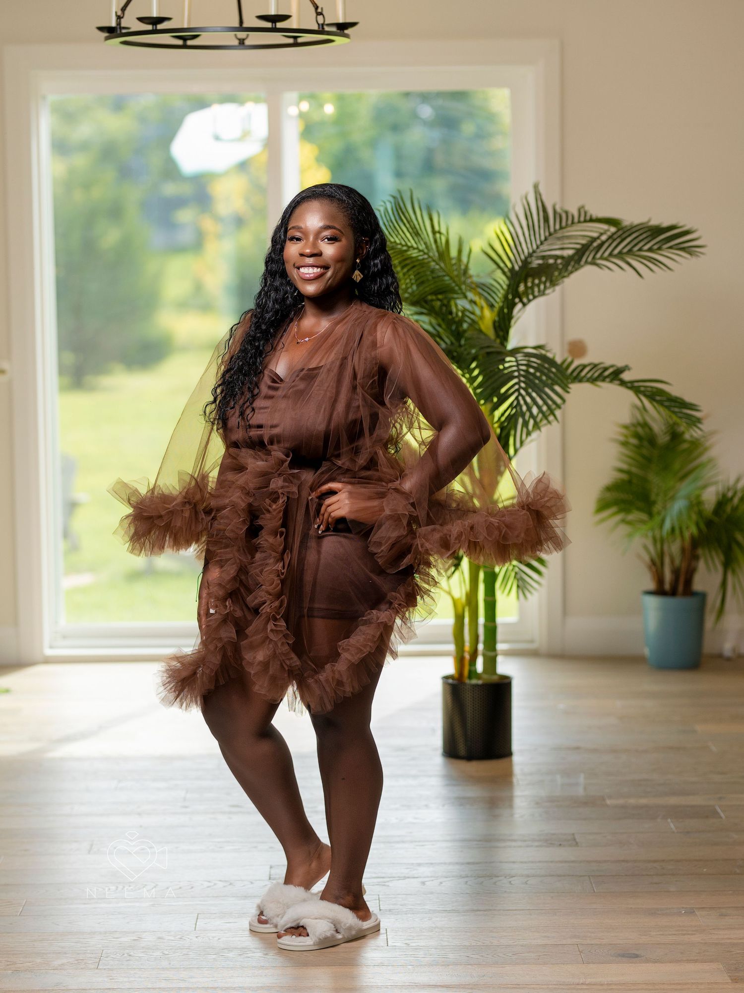 Woman in brown feathered loungewear poses playfully in bright interior with plants.