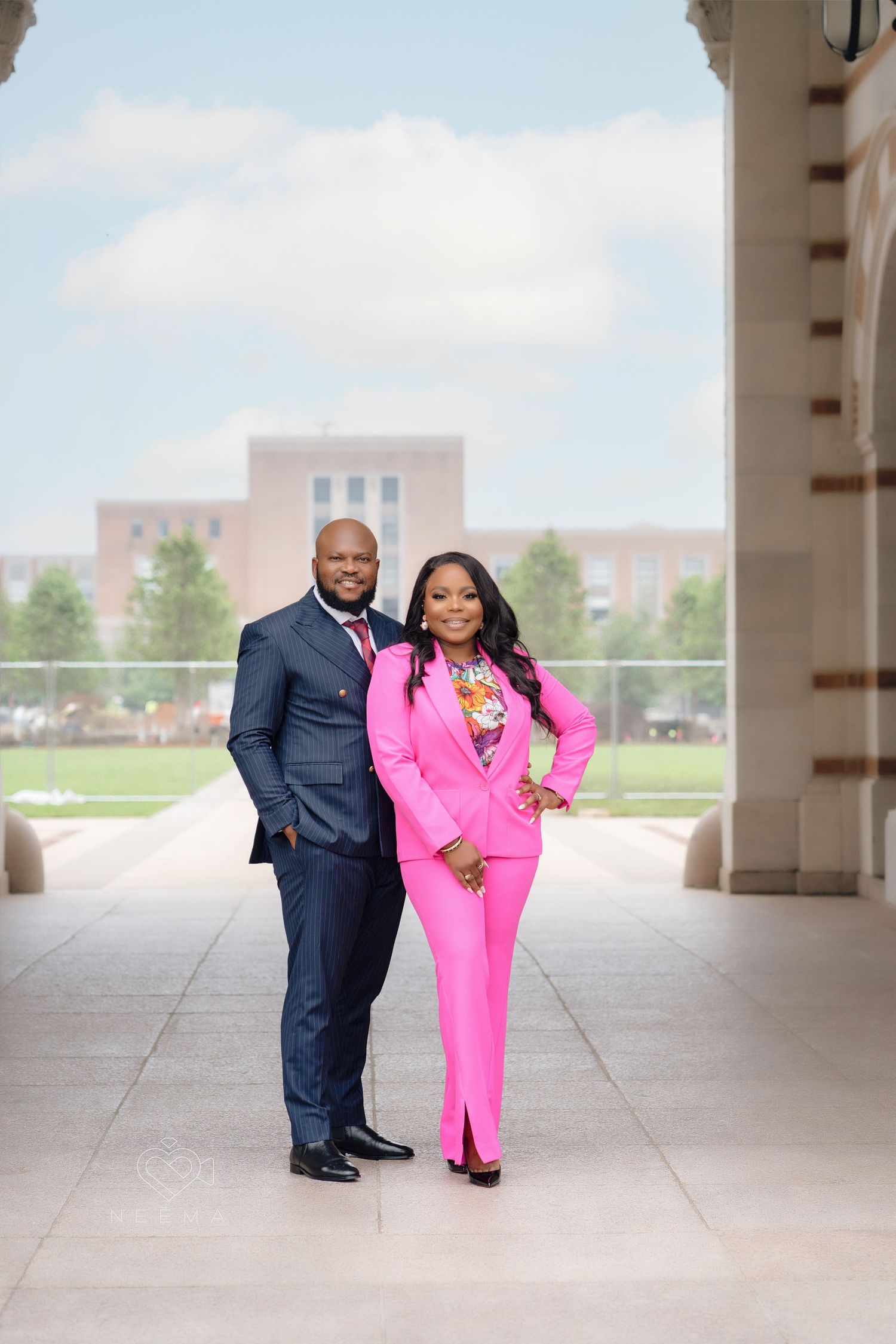 A couple in coordinated formal attire stands in a covered walkway with a navy suit and bright pink suit.