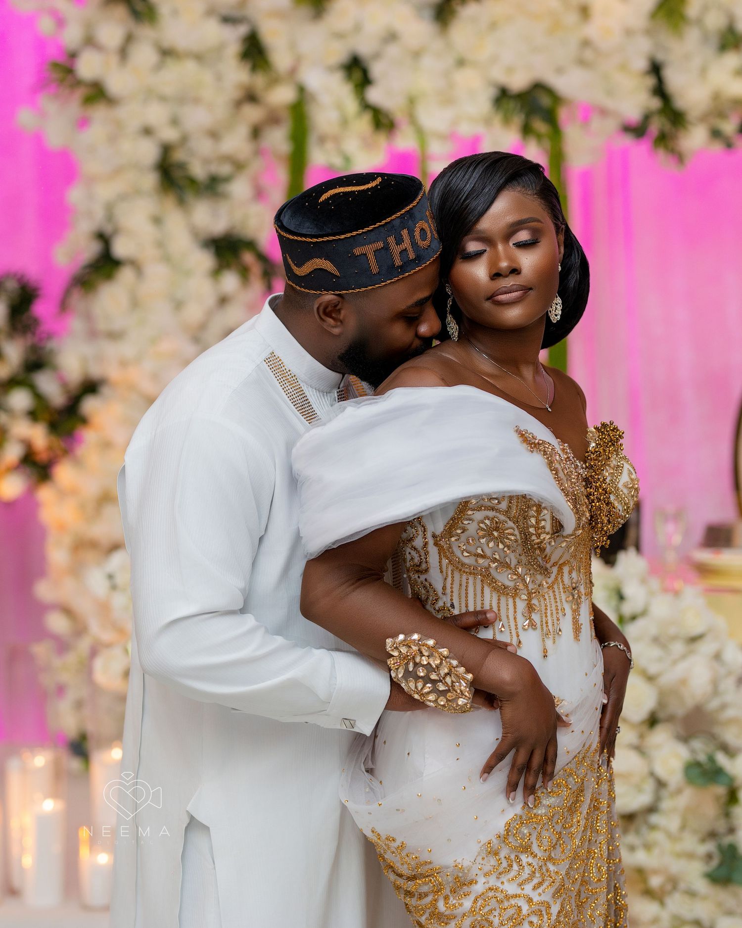 Romantic wedding portrait with gold and white formal attire against floral backdrop with pink lighting.