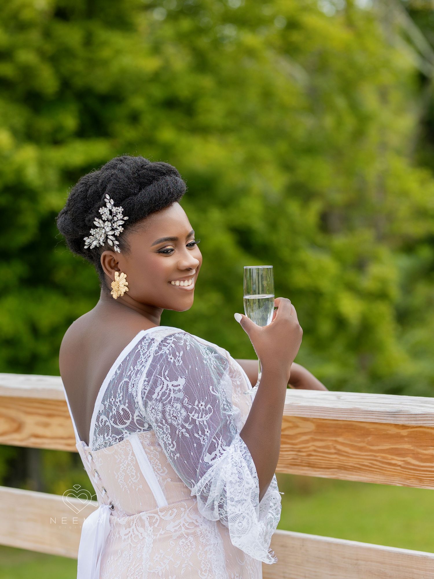 Bride in lace wedding dress holds champagne glass while smiling outdoors.