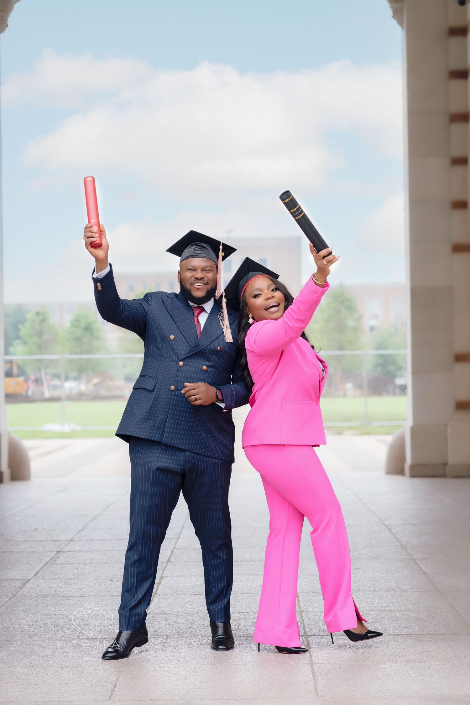 A celebrating couple in formal attire holds up graduation diplomas while posing under an archway.