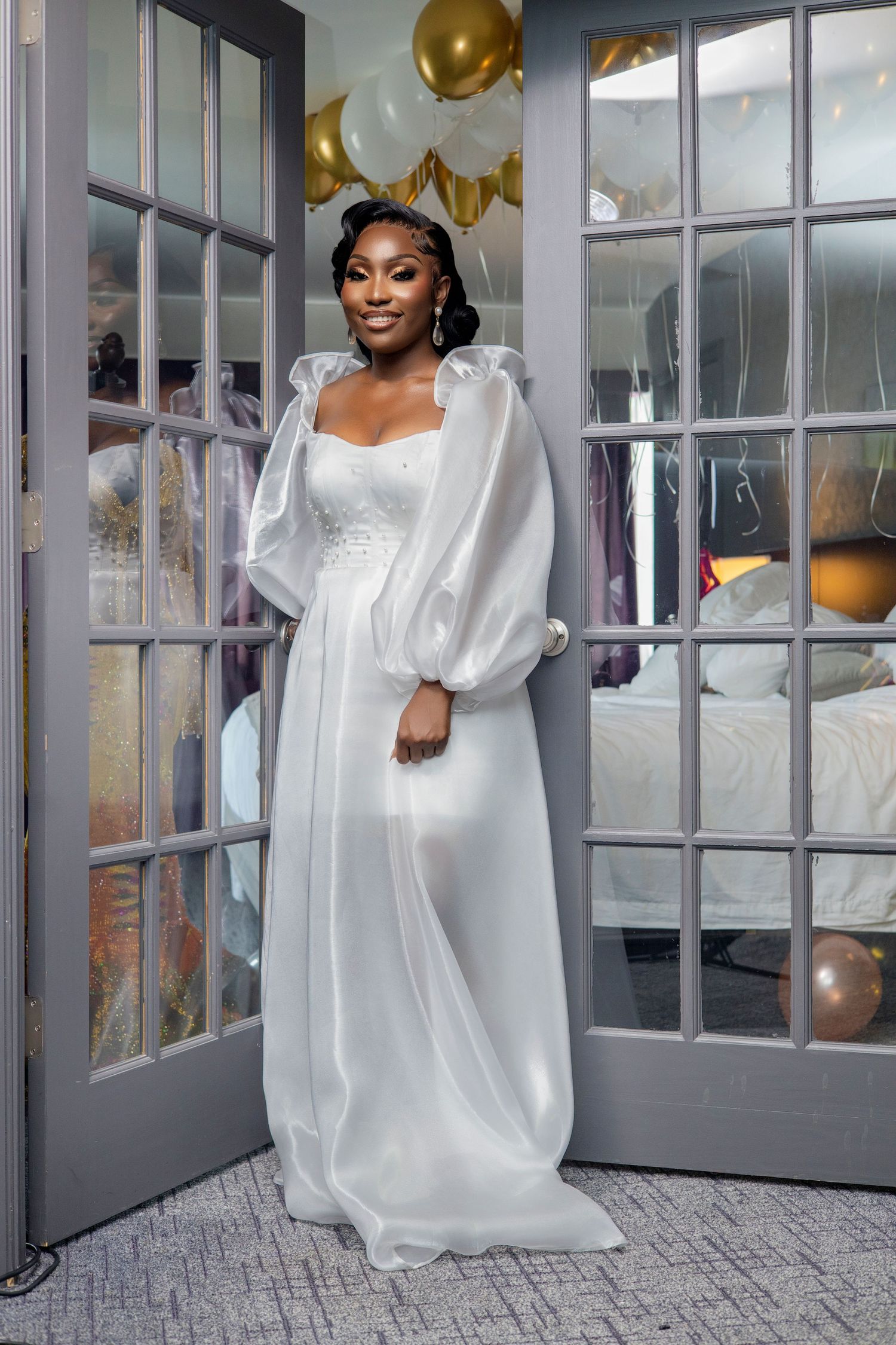 Elegant bride in flowing white gown with puffy sleeves poses by French doors with gold balloons in background.