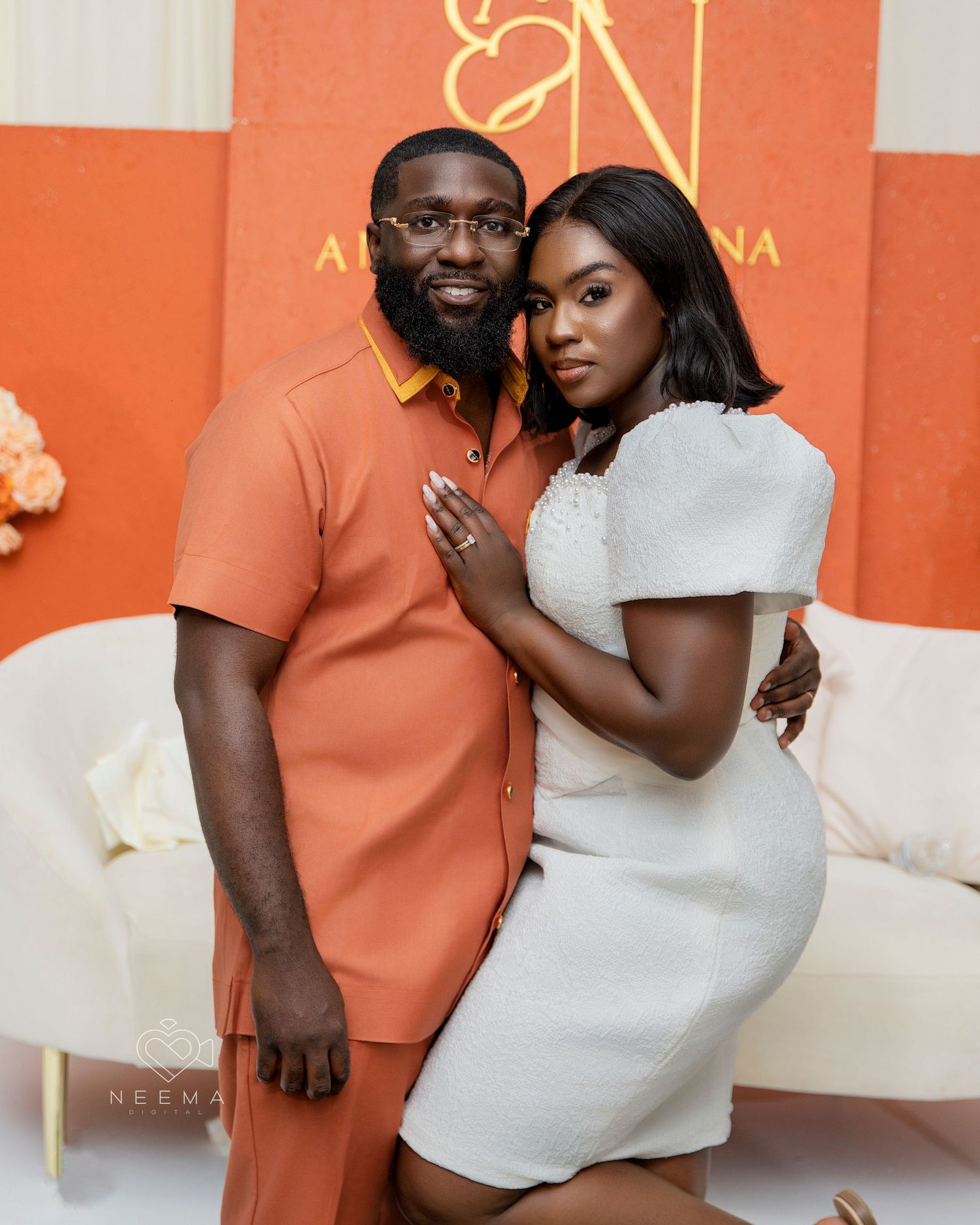 Couple in coordinating orange and white outfits pose against coral colored backdrop with floral decor.