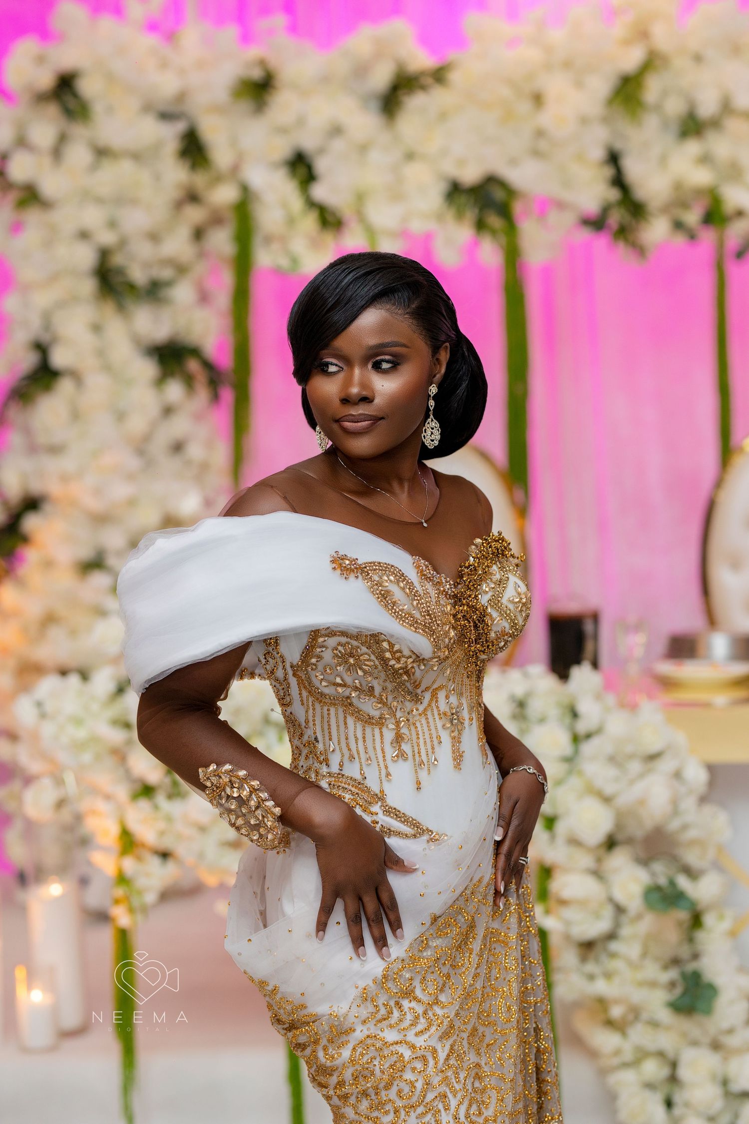 An elegant white and gold beaded gown with off-shoulder design against a pink and floral backdrop.