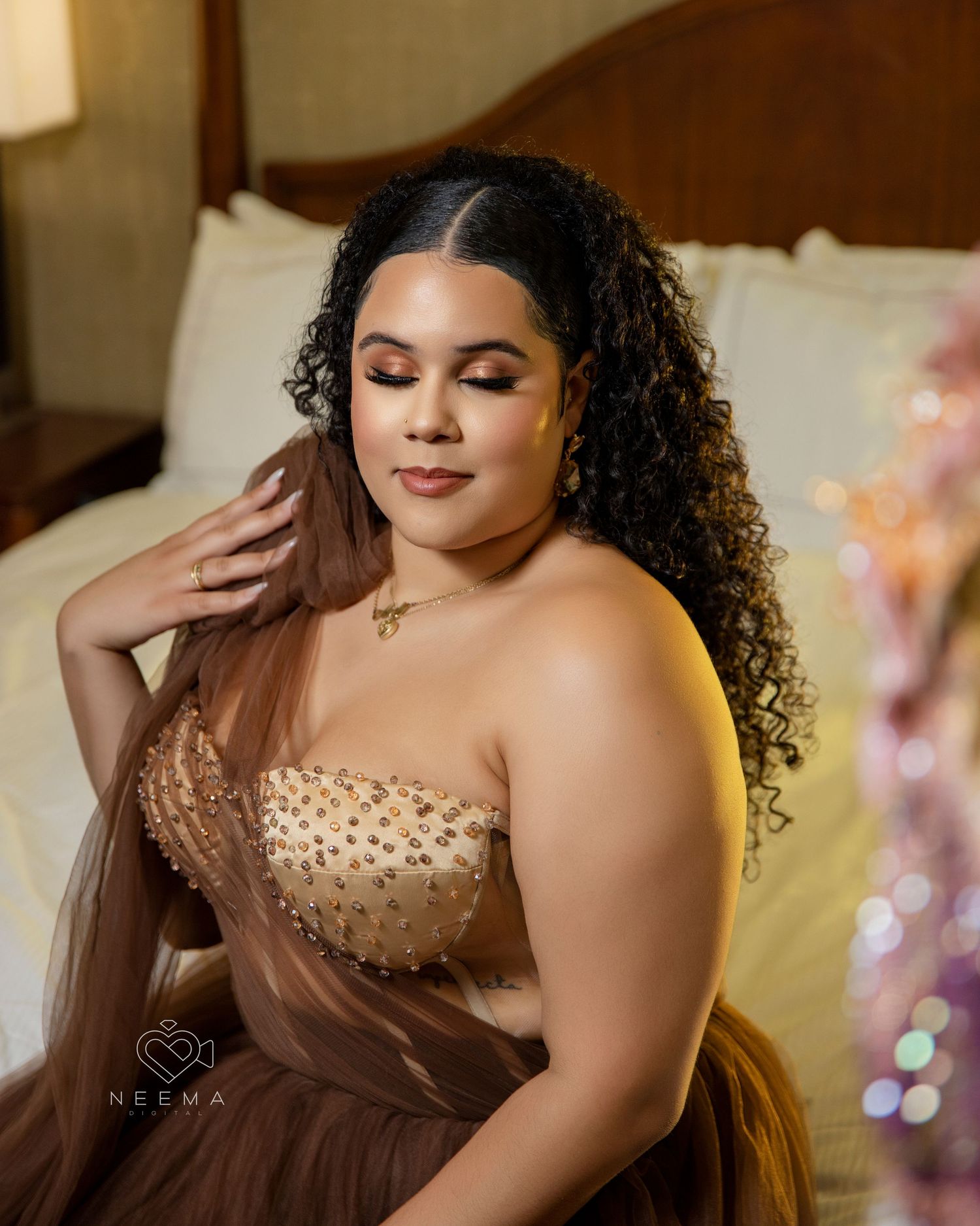 Person in brown tulle gown poses elegantly on bed with soft lighting.
