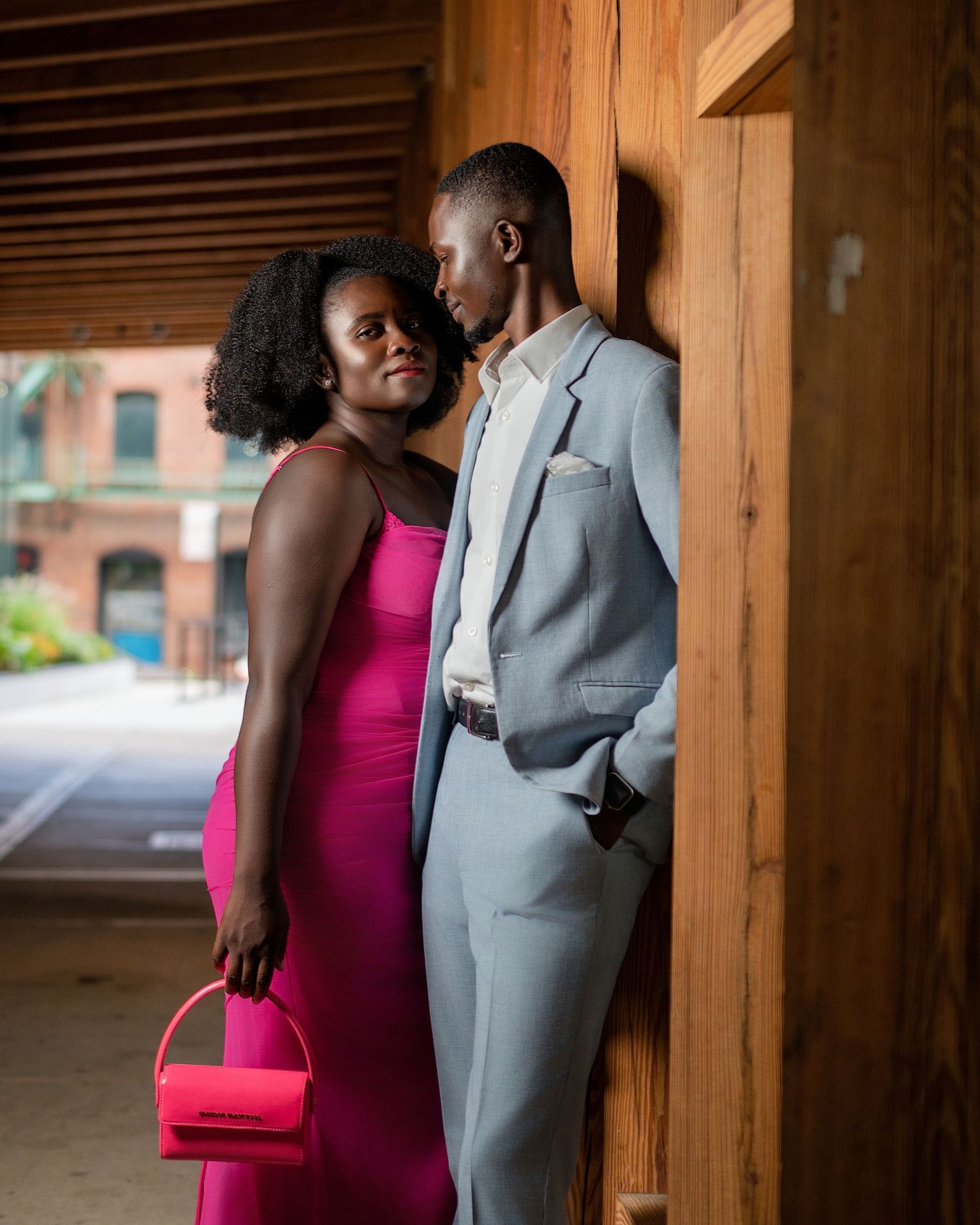Stylish couple in pink dress and grey suit pose against rustic wooden backdrop.