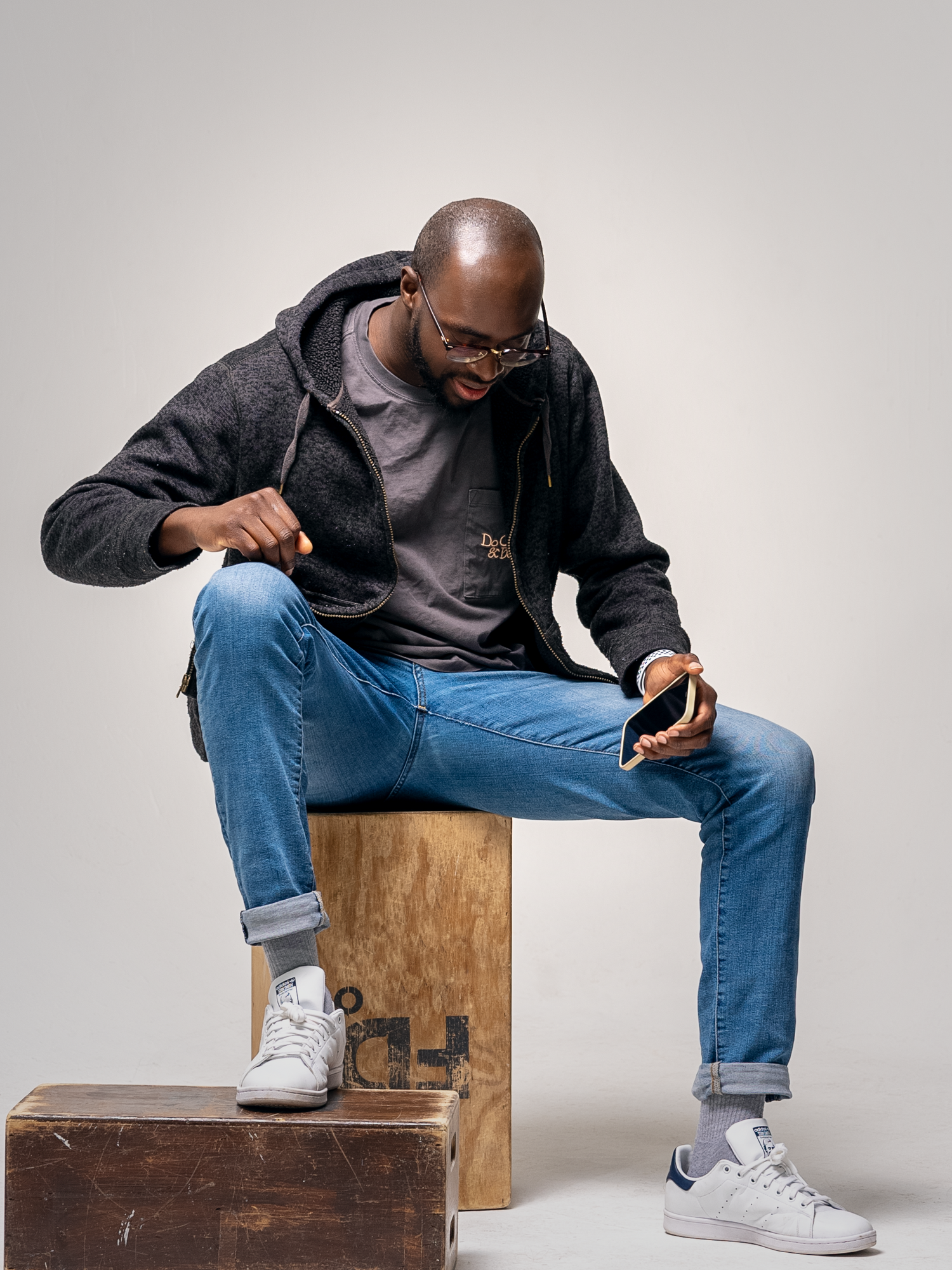 Casual portrait of a person in denim and hoodie sitting on wooden crates in studio setting.