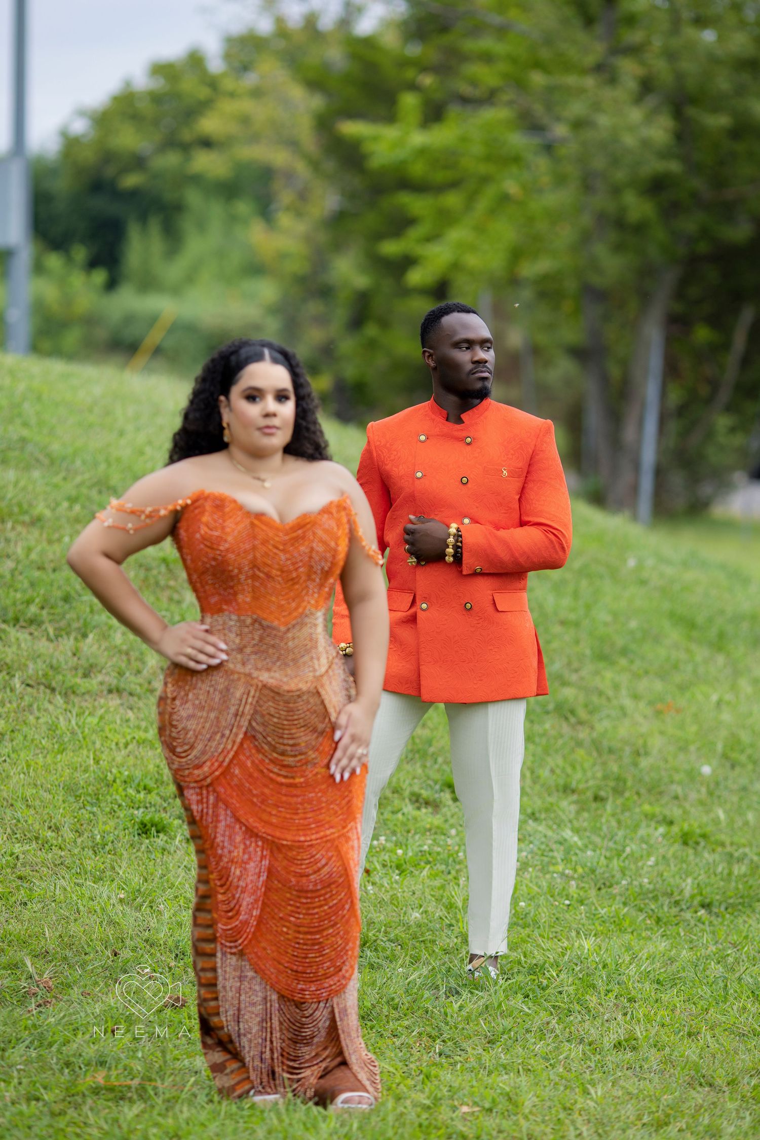 A couple poses outdoors wearing formal attire in orange and coral tones.