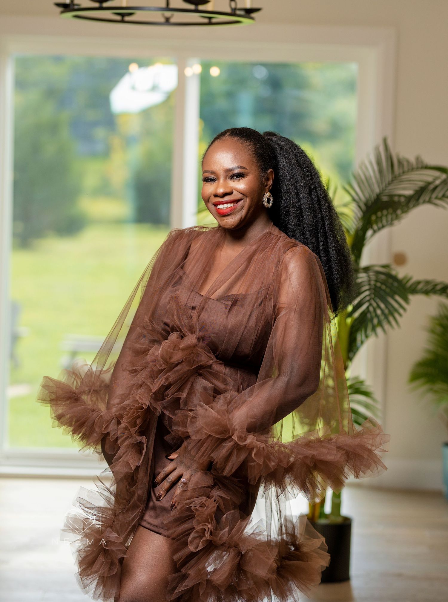 A woman in a dramatic brown tulle gown poses elegantly near a window with natural lighting.