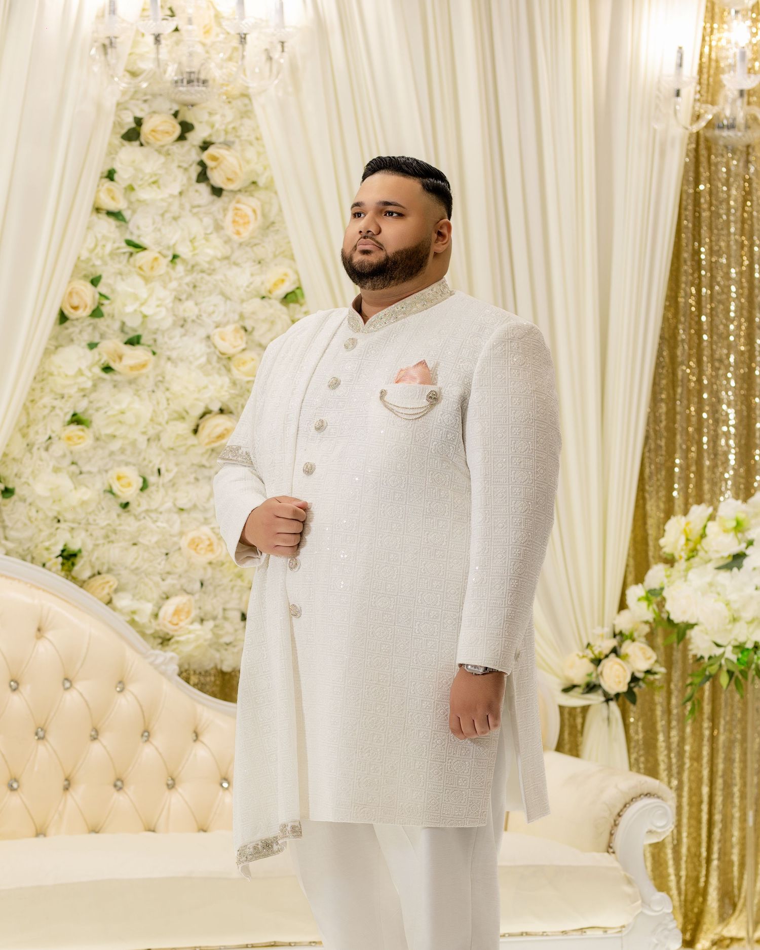 Groom in classic cream Nehru-collar suit stands against floral wall backdrop.