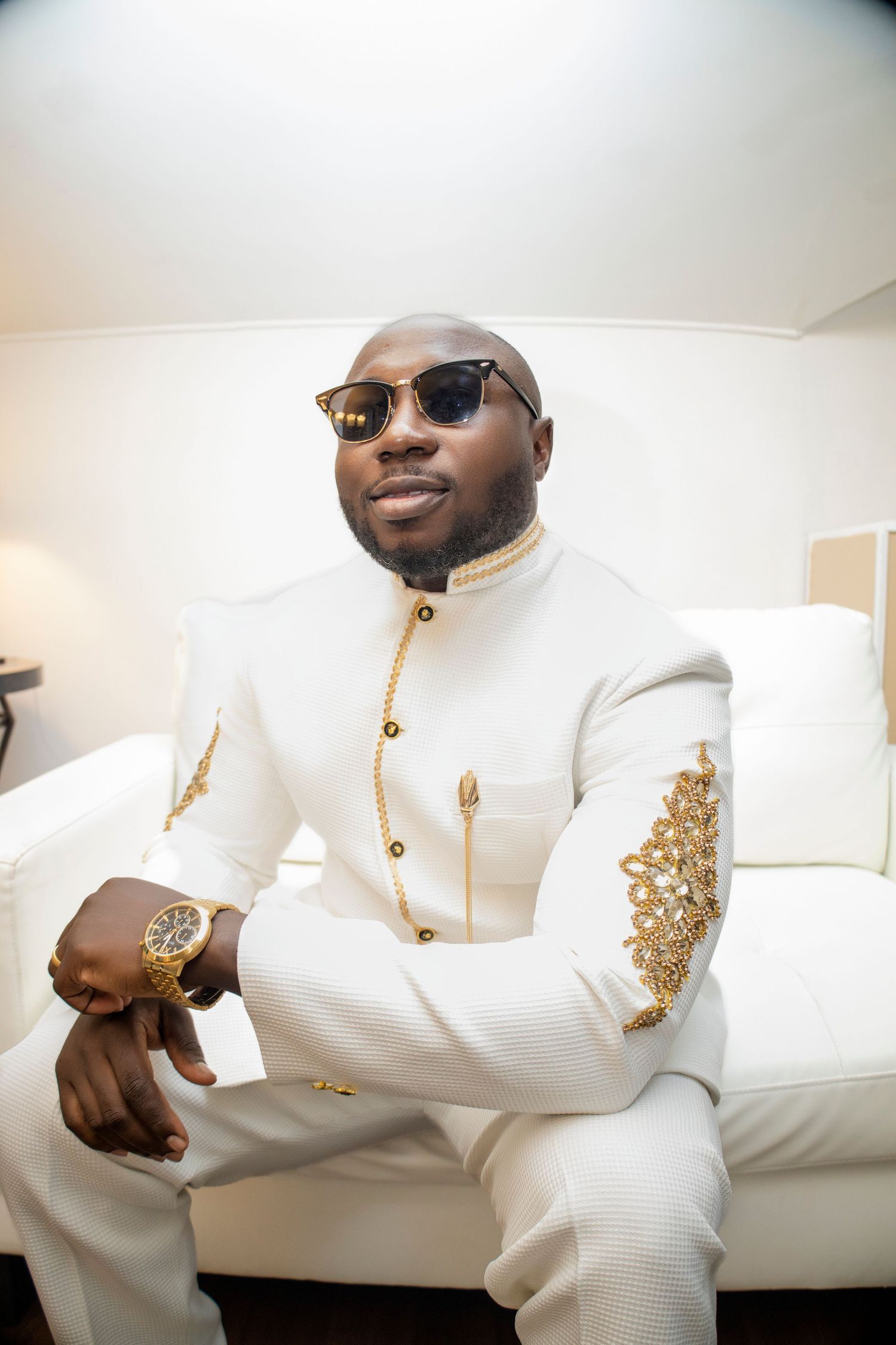 Stylish groom in white embroidered suit and sunglasses poses confidently on couch.
