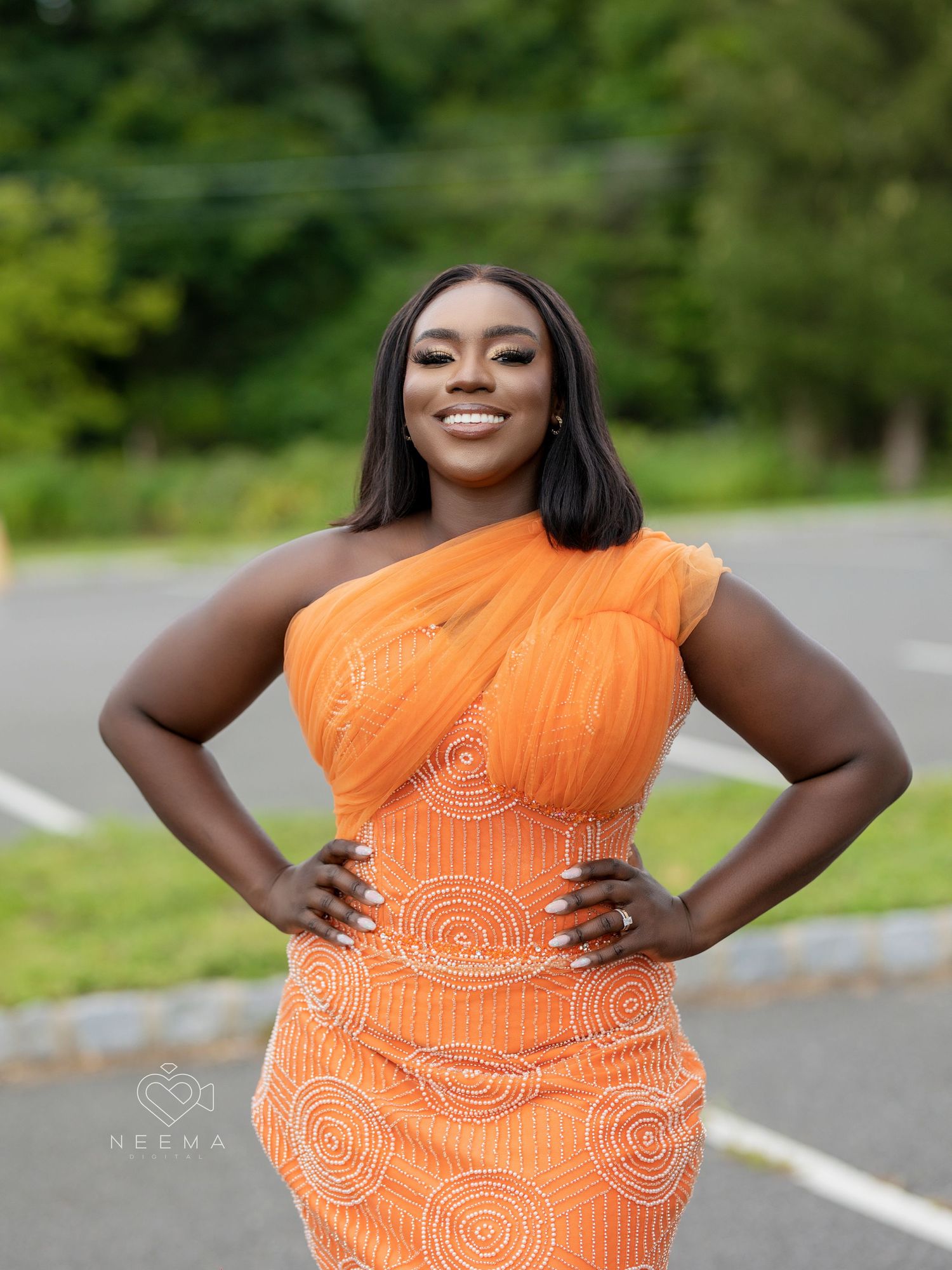 Woman wearing vibrant orange one-shoulder dress with circular pattern stands confidently on outdoor path.