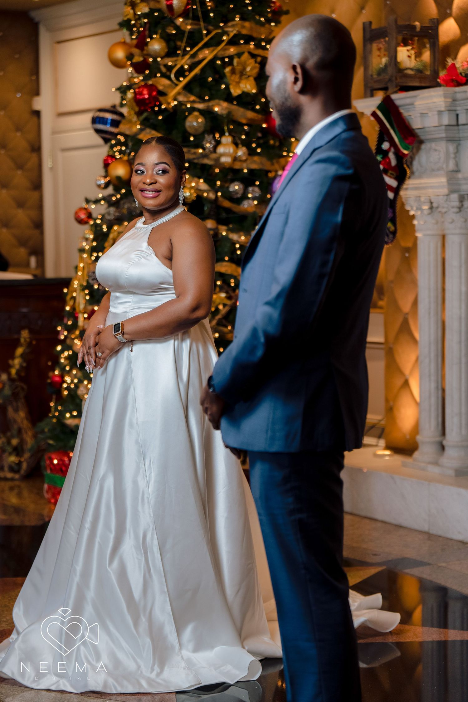 Couple in wedding attire poses by decorated Christmas tree and fireplace.