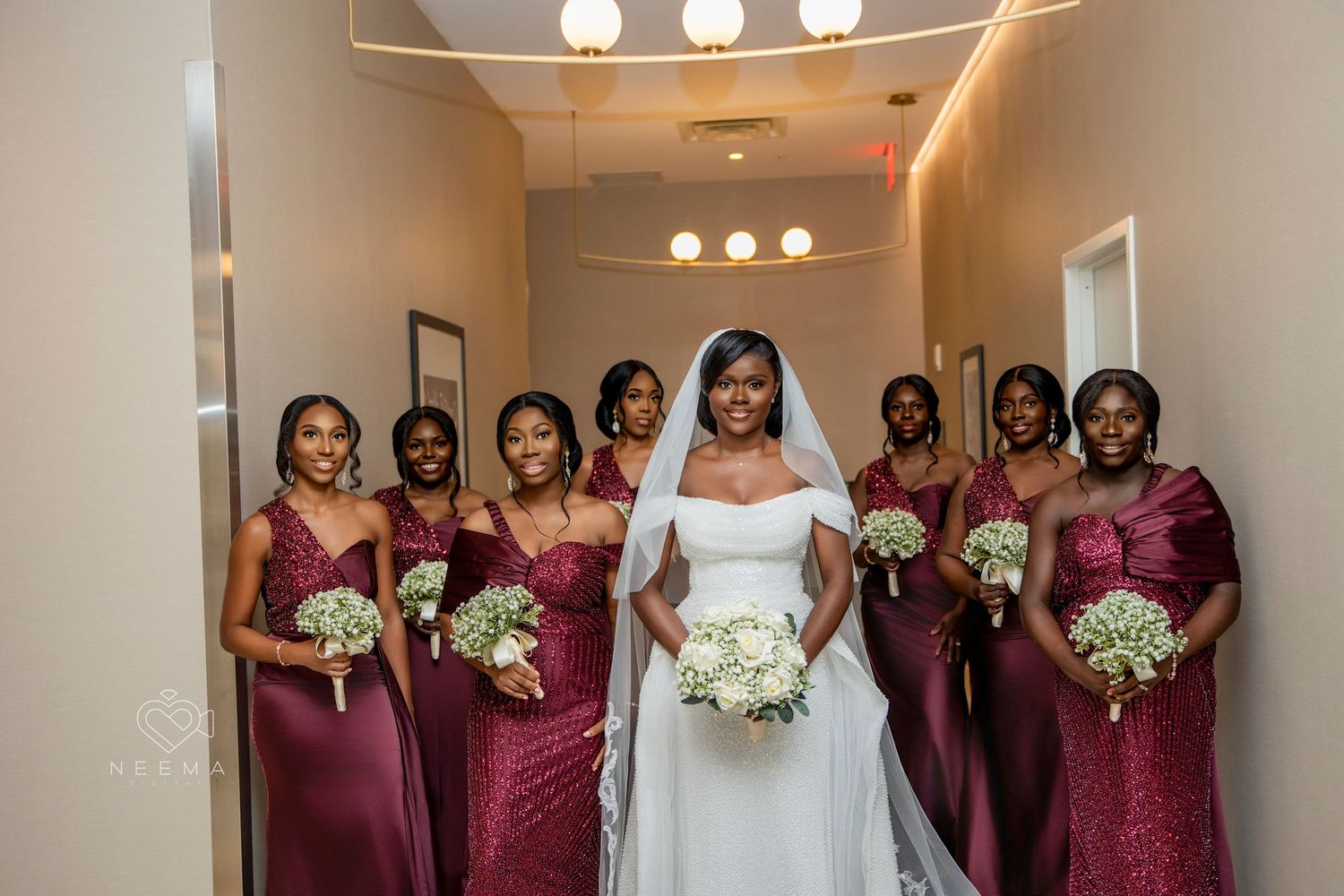 Bridal party in burgundy dresses poses with bride holding white bouquets in hallway.