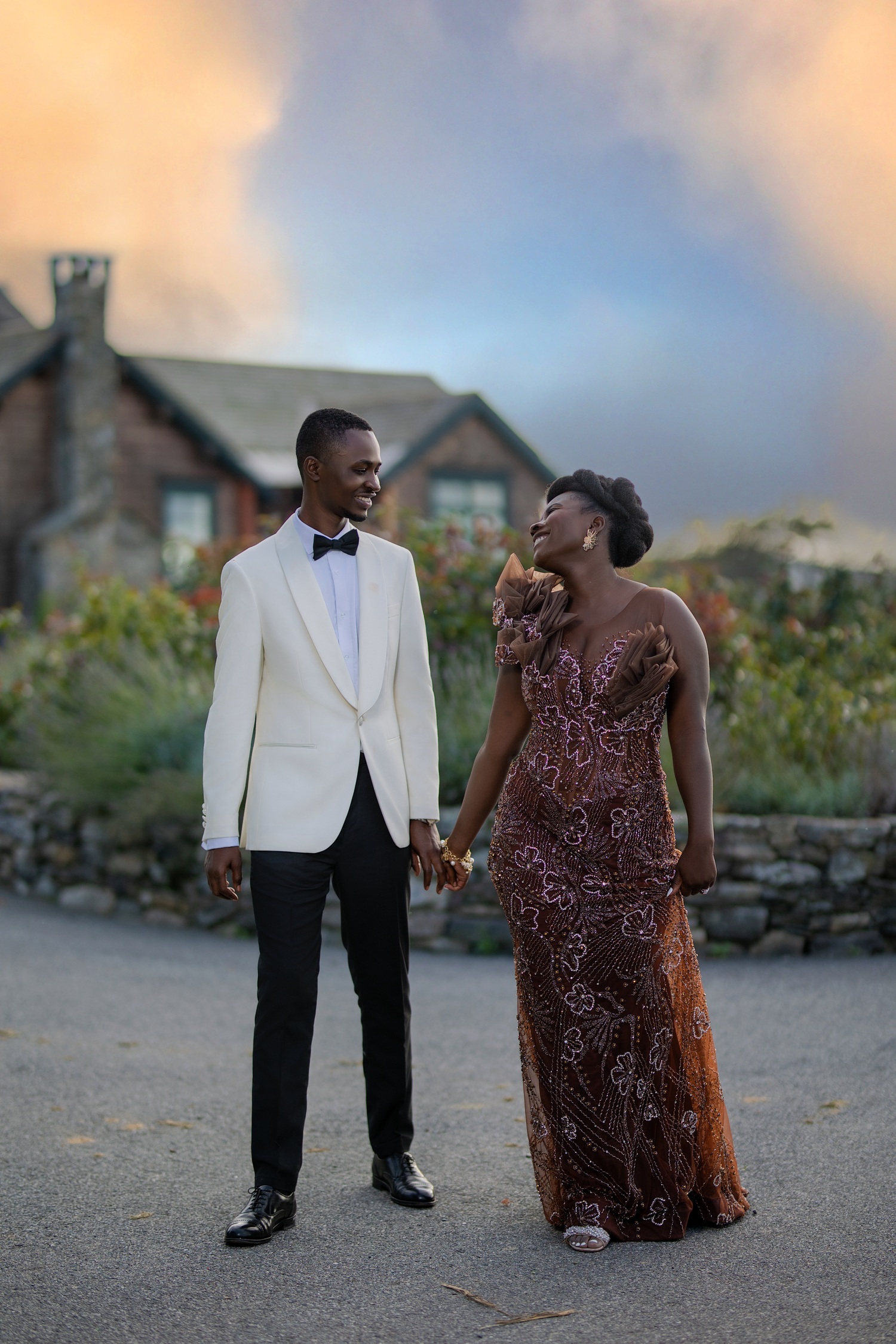 A couple shares a joyful moment in formal attire against a stone wall and sunset backdrop.