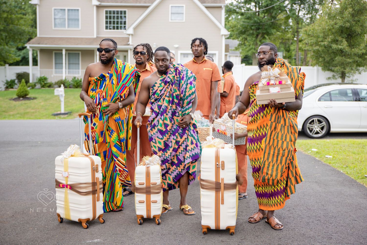 Group wearing vibrant African print outfits pose with matching luggage on residential street.