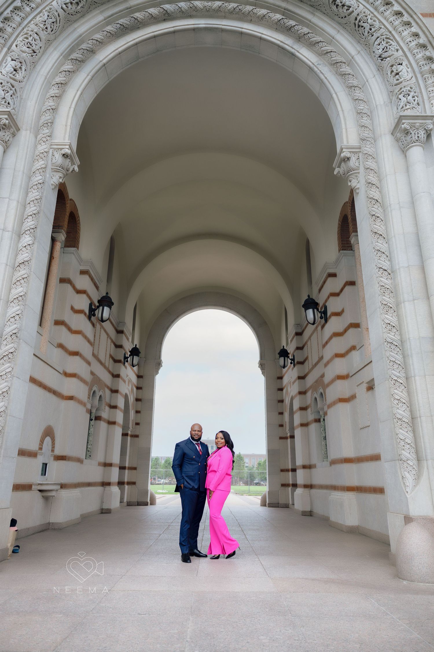 A couple poses together under an ornate white archway in formal attire with a scenic background.