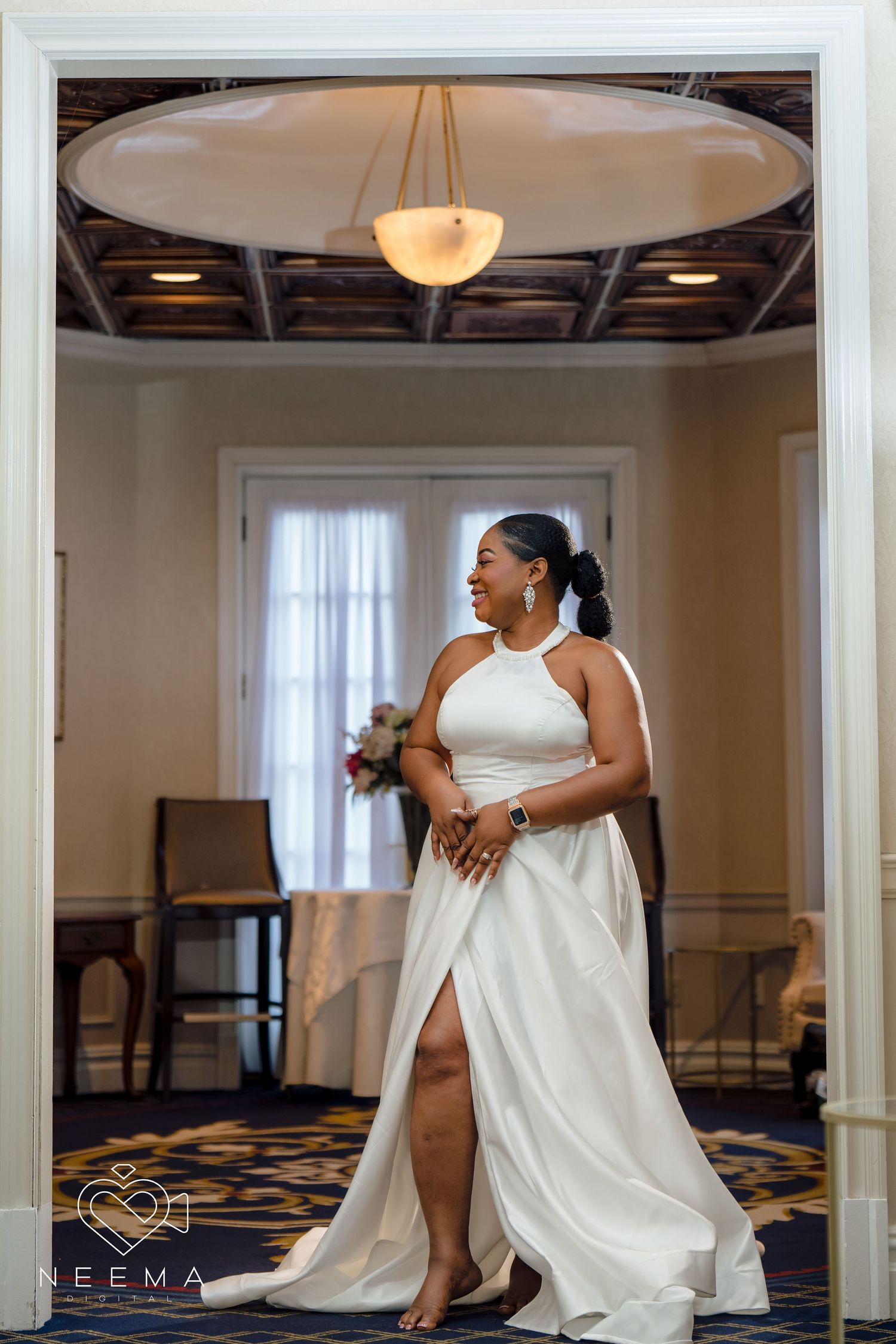 Elegant bride in white halter dress poses beneath ornate chandelier in luxurious venue.