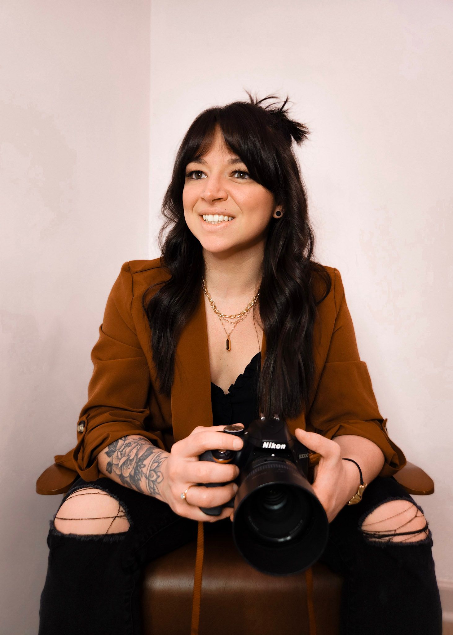 Emily Hall, photographer in a brown blazer and ripped jeans sits with a Nikon camera, showing visible arm tattoos.