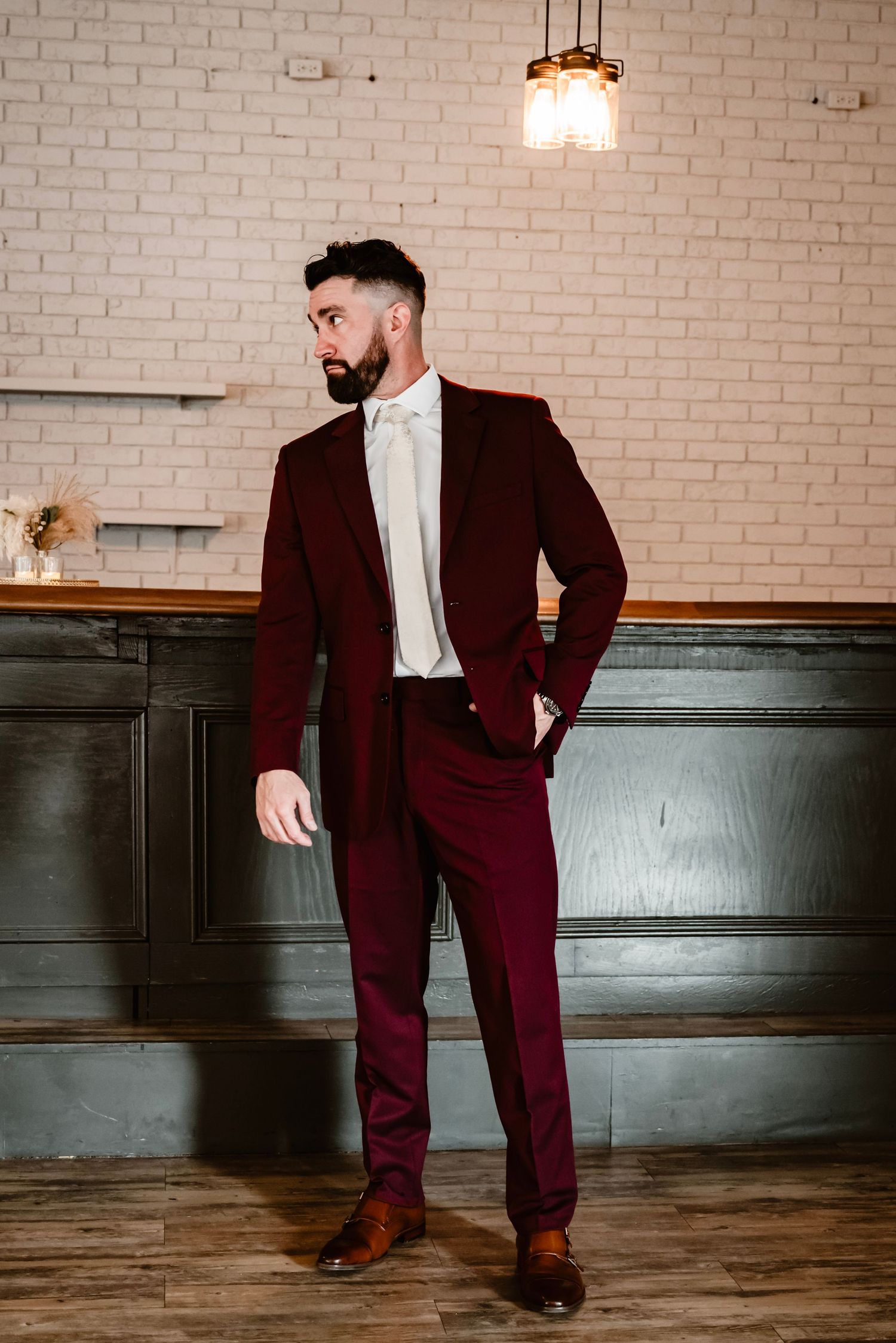 A groom in a burgundy suit and white shirt poses against a brick wall at The Little Cottage in Smyrna, Georgia