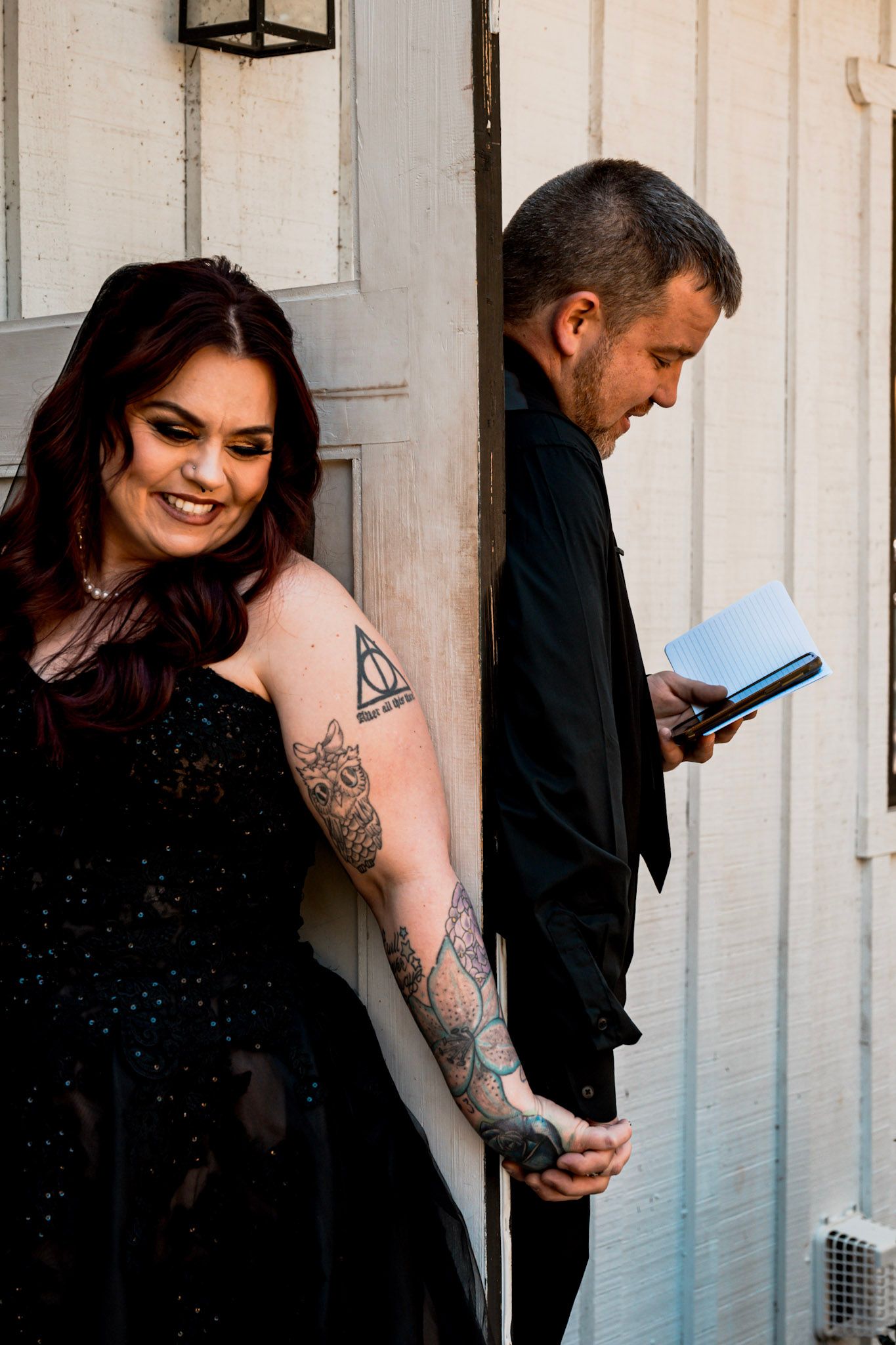 A couple stands by a door, with the bide in a black dress showing tattoos while the groom  reads their vows at Koury Farms in Auburn, GA.