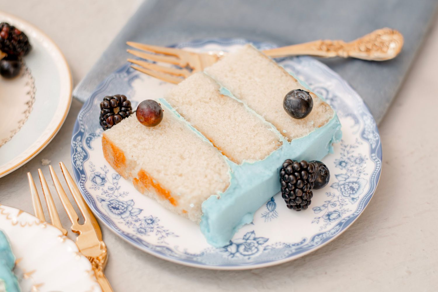A slice of layered cake with blue frosting served on a decorative plate with fresh blackberries and golden utensils.