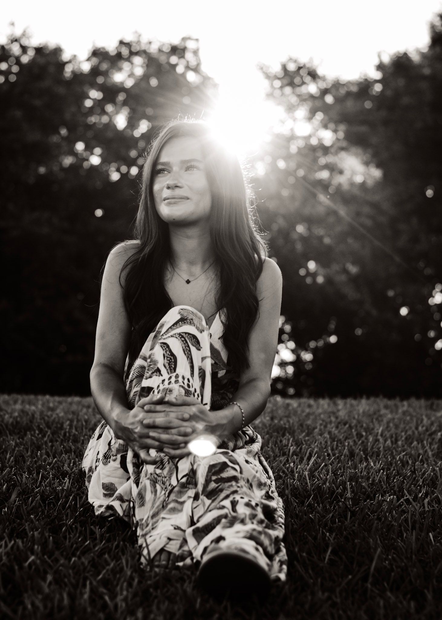 A high-school senior in a floral dress sits in sunlit grass creating a dreamy black and white portrait in Covington, georgia.