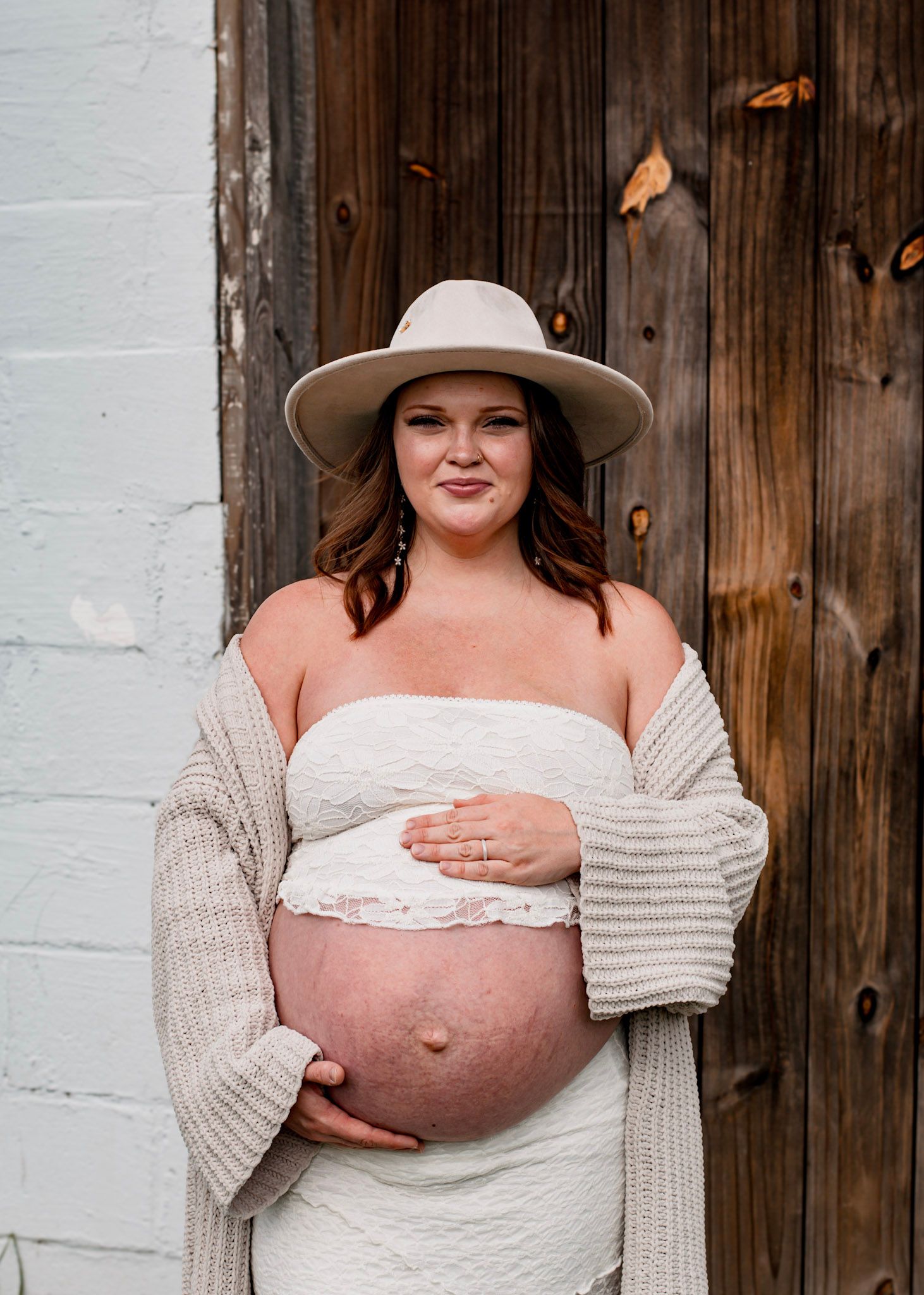 Pregnant woman in beige sweater and hat poses against rustic wooden door in maternity photo shoot.