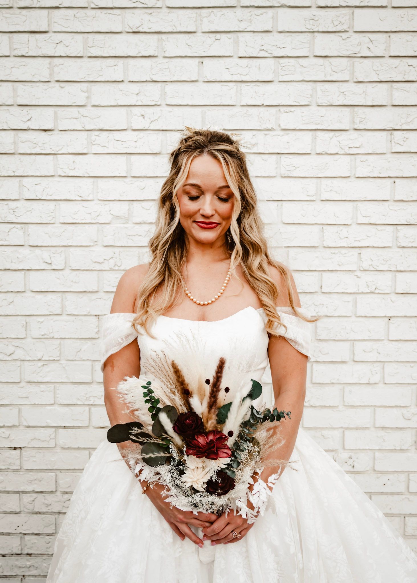 Bride in strapless white lace wedding gown holds burgundy and white floral bouquet against white brick wall The Little Cottage in Smyrna, GA