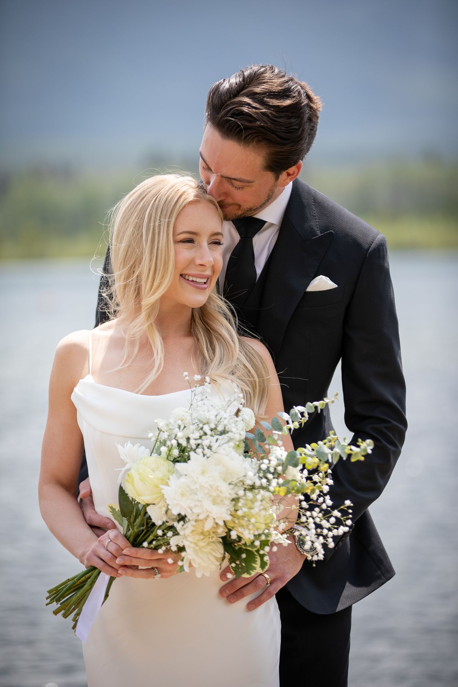 A couple in wedding attire shares intimate moments by Vermilion lake with white floral bouquets in a series of romantic photos. Banff, AB